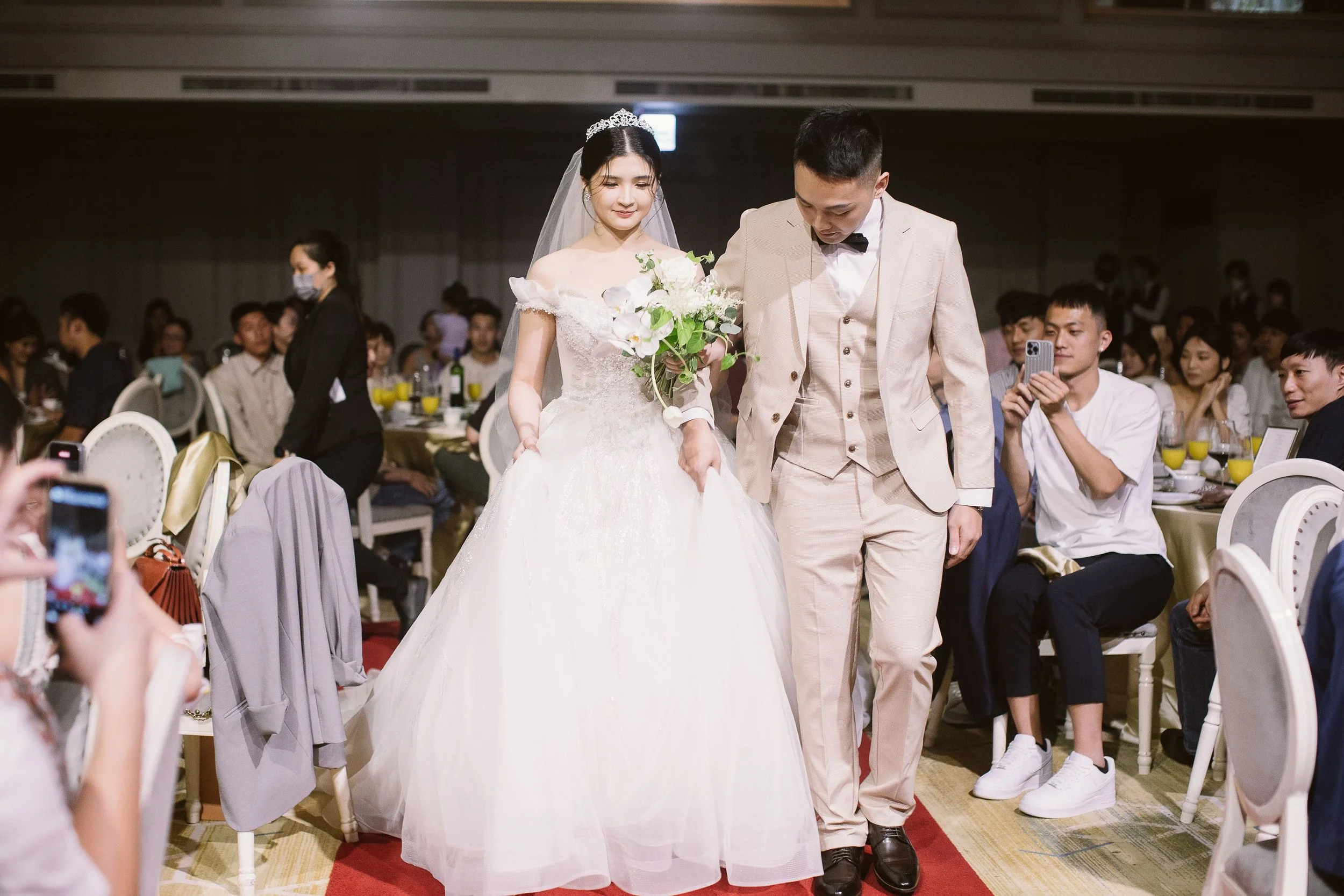 A bride and groom walking together in a wedding reception hall, surrounded by seated guests. The bride is wearing a white wedding gown with lace details and a tiara, holding a bouquet, while the groom is in a light-colored suit with a black bow tie. 