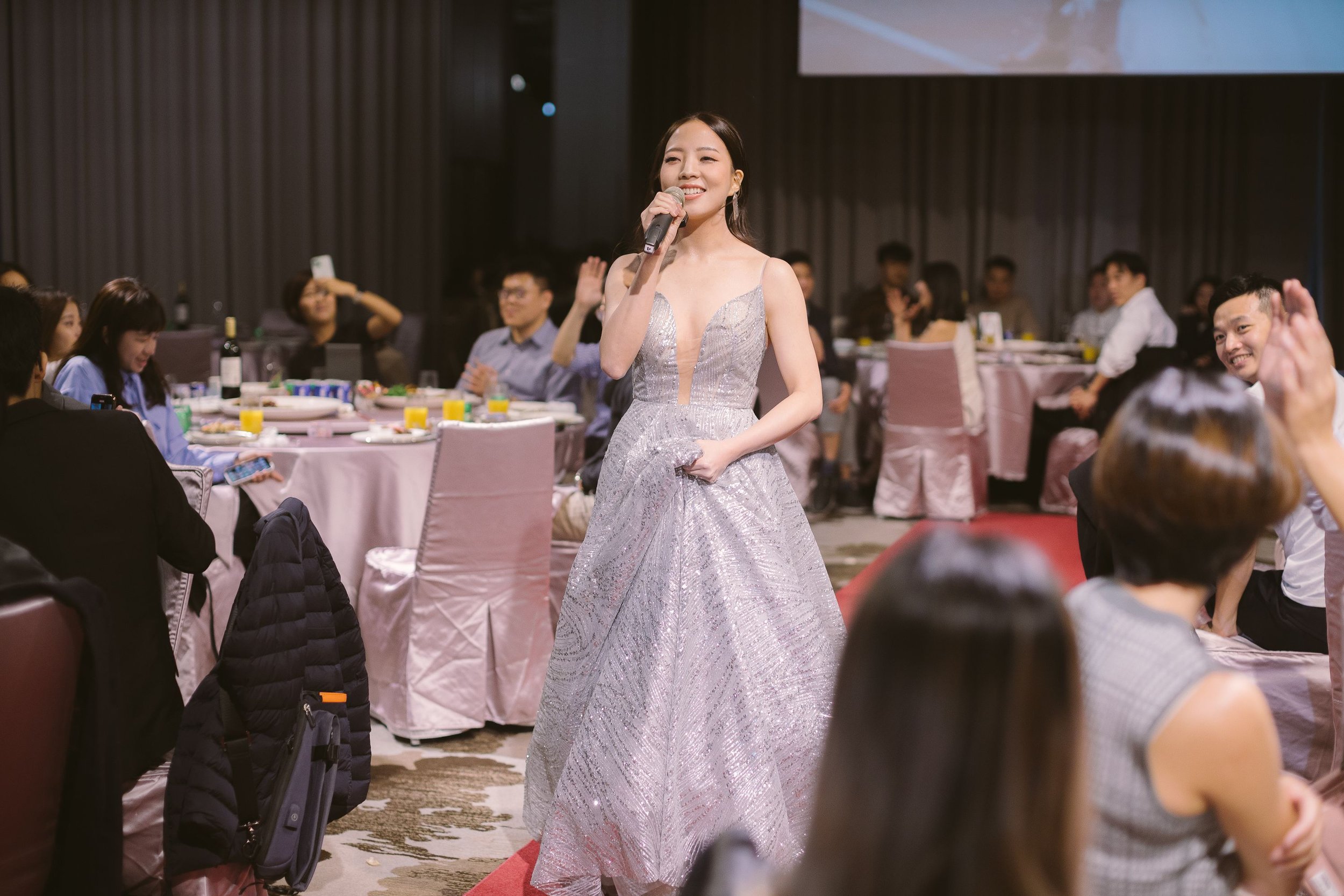 A woman in a silver glittery gown is smiling and holding a microphone at a social event with several people seated at dinner tables in the background.