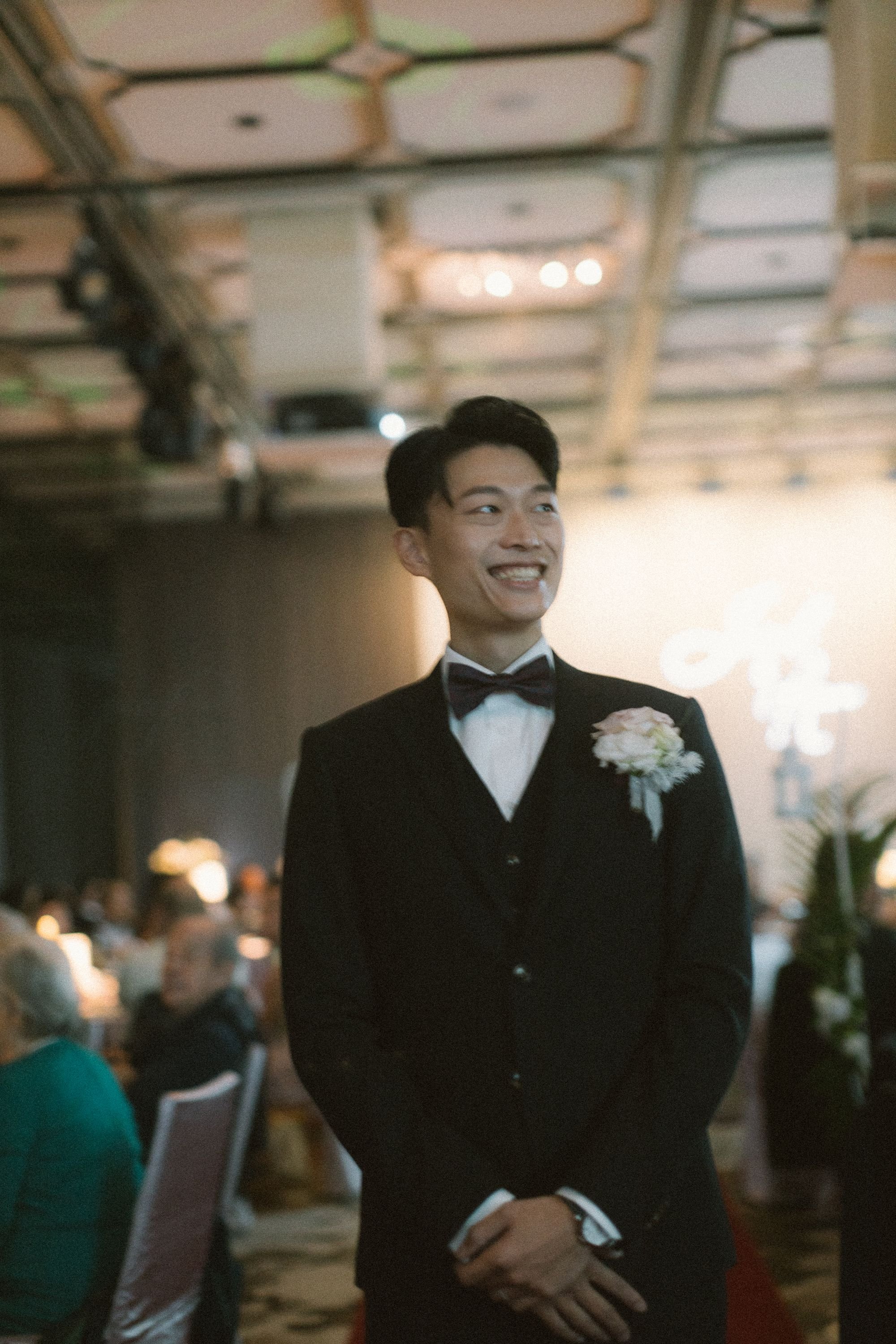 A young man dressed in a black tuxedo with a white shirt and a bow tie, smiling and standing with his hands clasped together at a formal event or wedding reception.