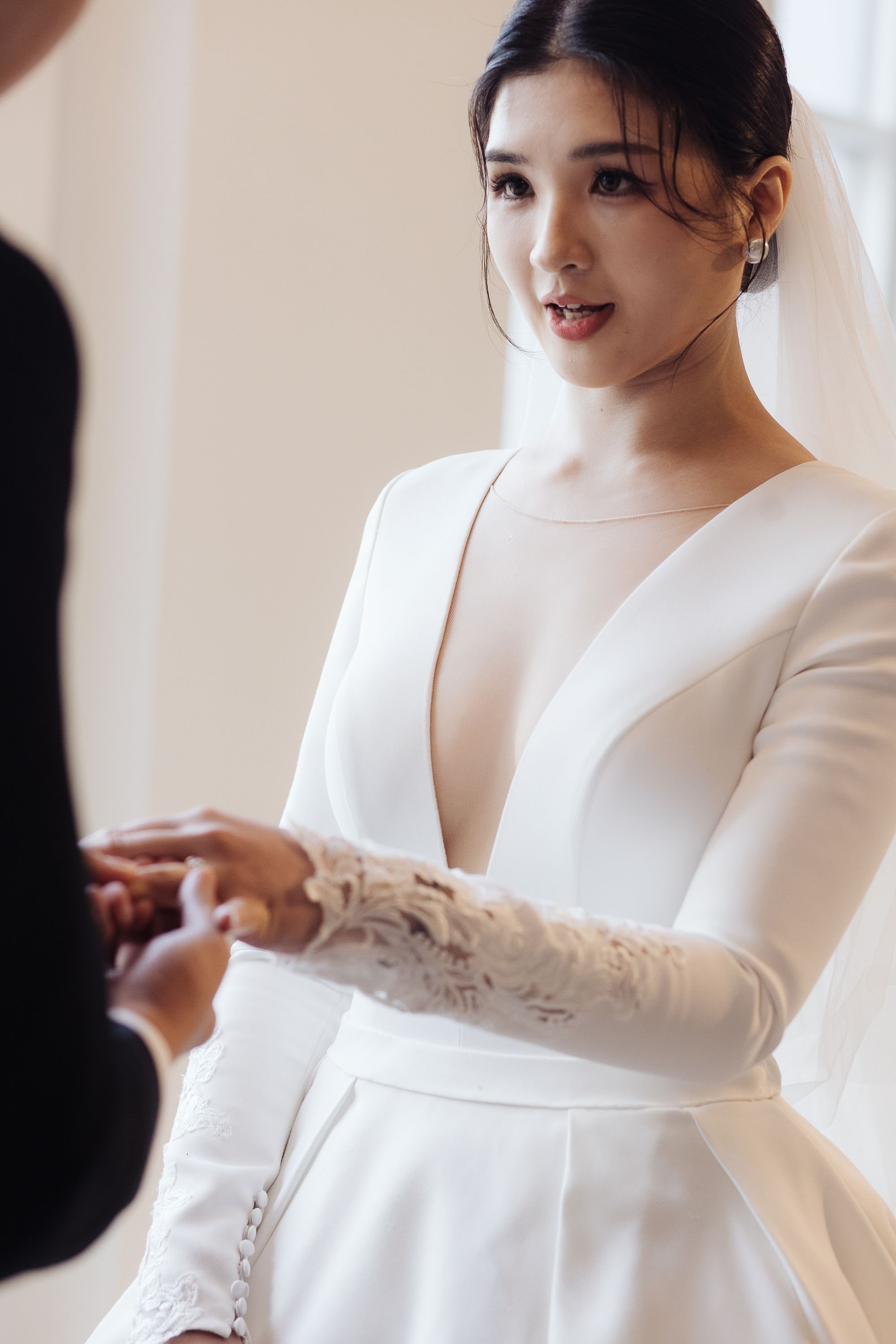 A bride in a white wedding dress with lace details on the sleeves and buttons on the wrist, smiling and holding hands with her partner during a wedding ceremony.