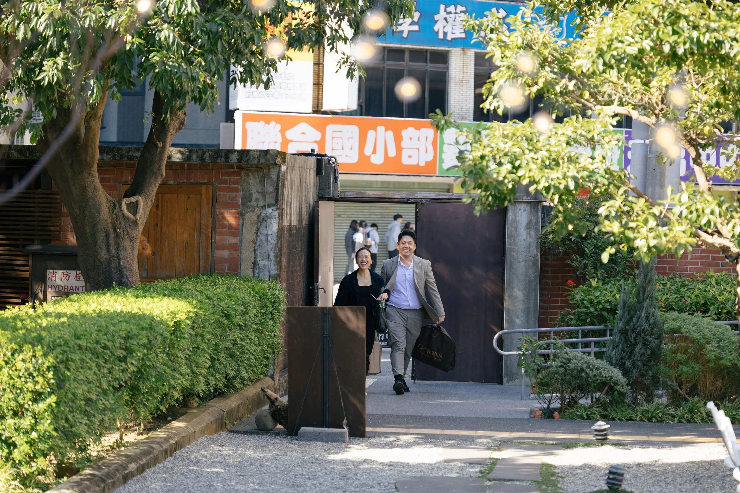 Two people, a man in a suit and a woman in a black jacket, walking out of a gated entrance in an urban park, surrounded by greenery and trees, with signs and buildings in the background.