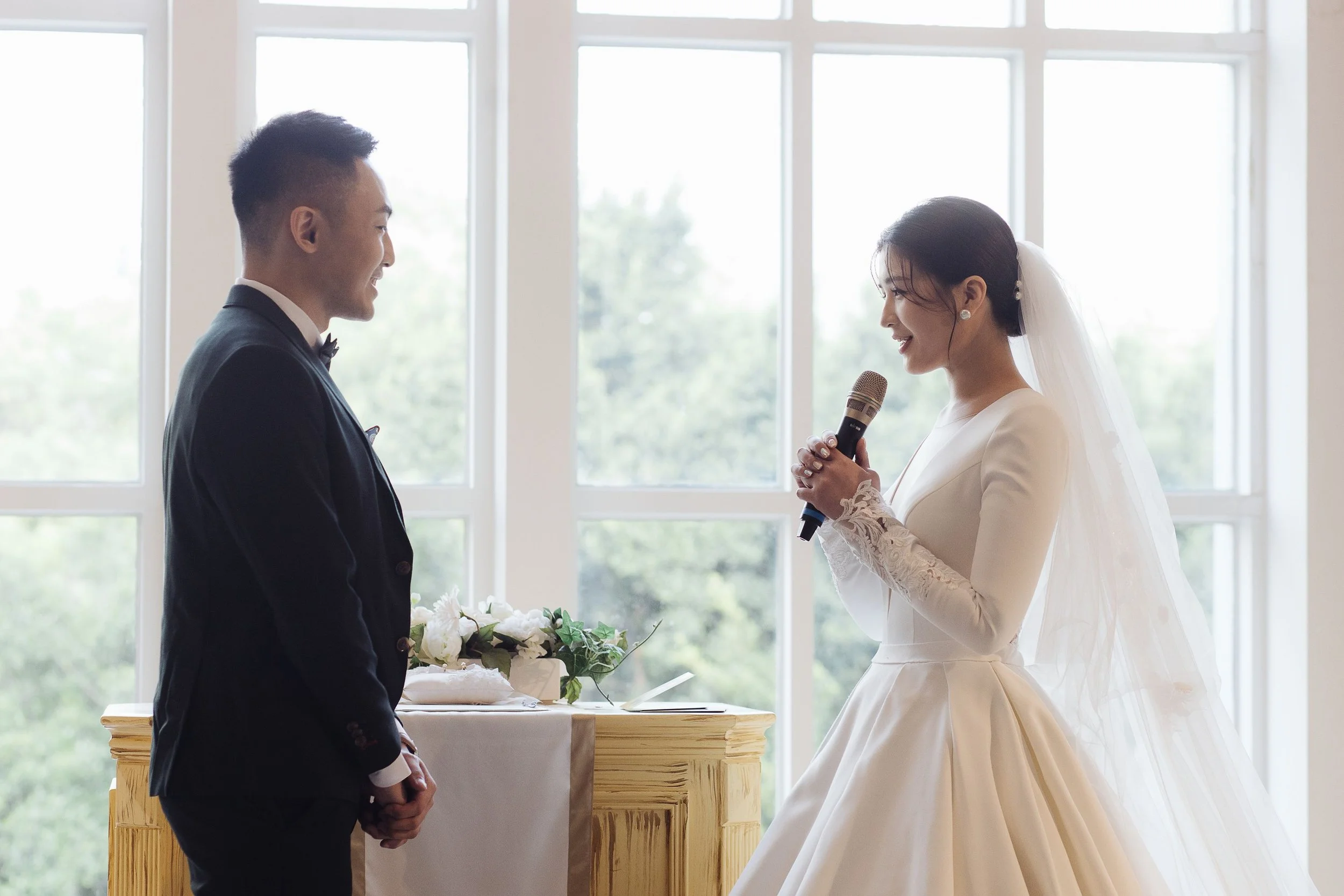 A bride and groom during their wedding ceremony in front of large windows, with the bride holding a microphone and the groom smiling.