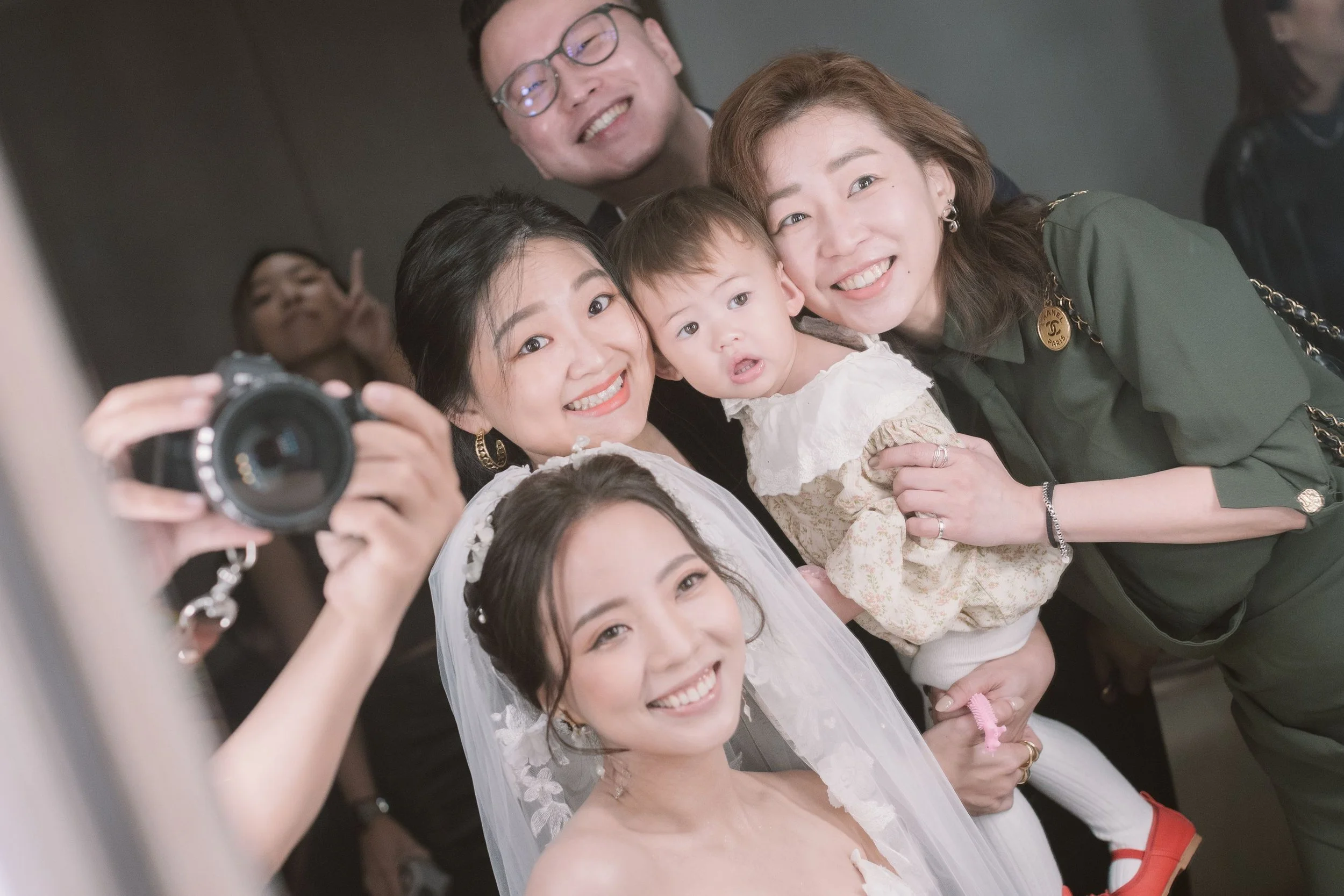 A group of smiling people taking a selfie, including a woman with a white veil, a small child, two women, and a man with glasses in the background, all smiling and looking at the camera.