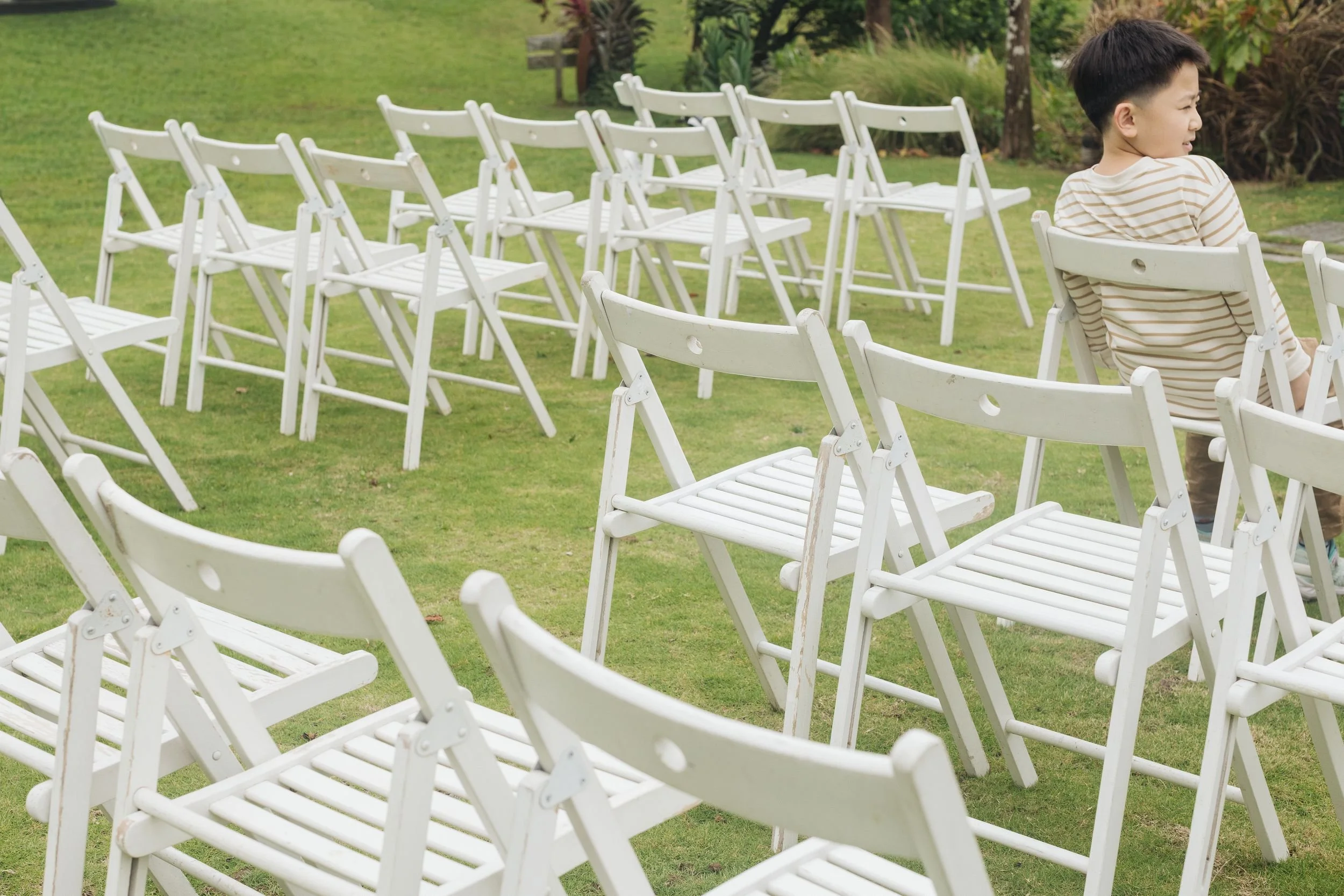 A young boy sitting on a white chair in an outdoor garden setting, with additional white chairs arranged nearby on a grassy area, surrounded by trees and plants.