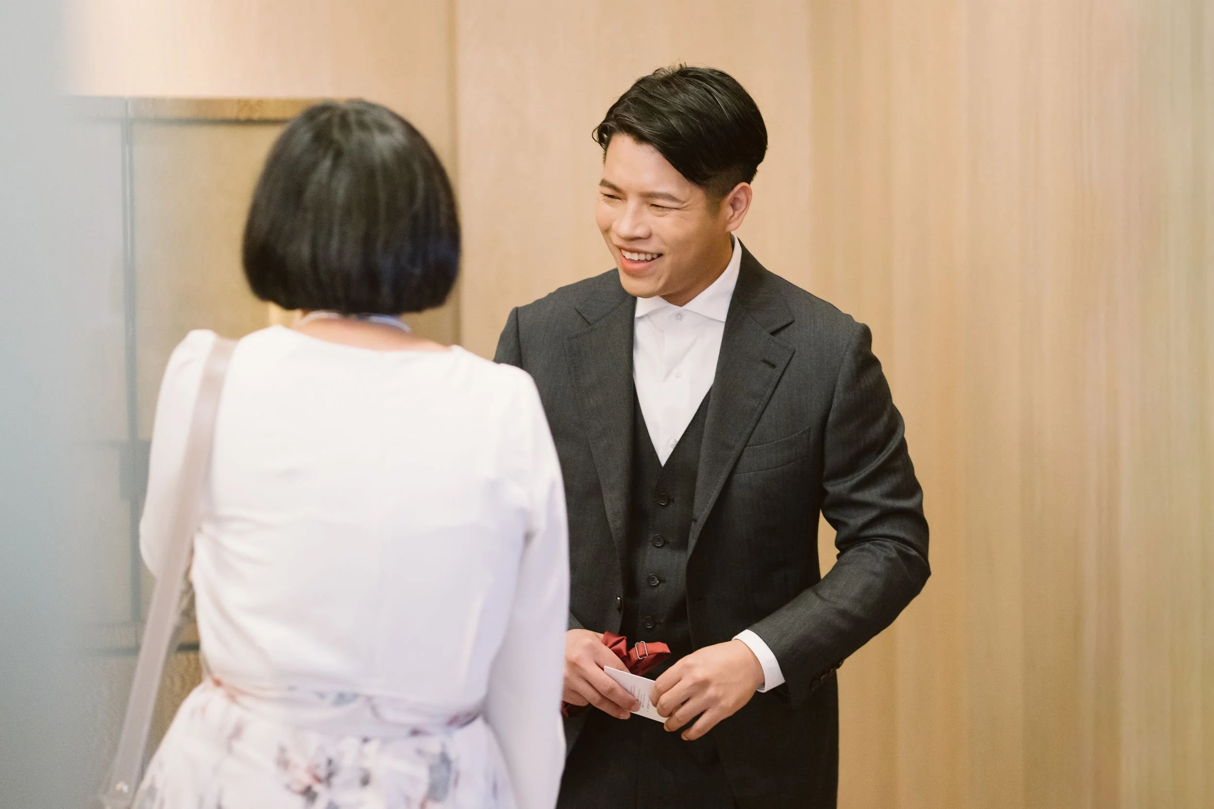 A man dressed in a dark suit and white shirt is smiling and talking to a woman with short dark hair, dressed in light-colored clothing, in a professional setting with wooden walls.