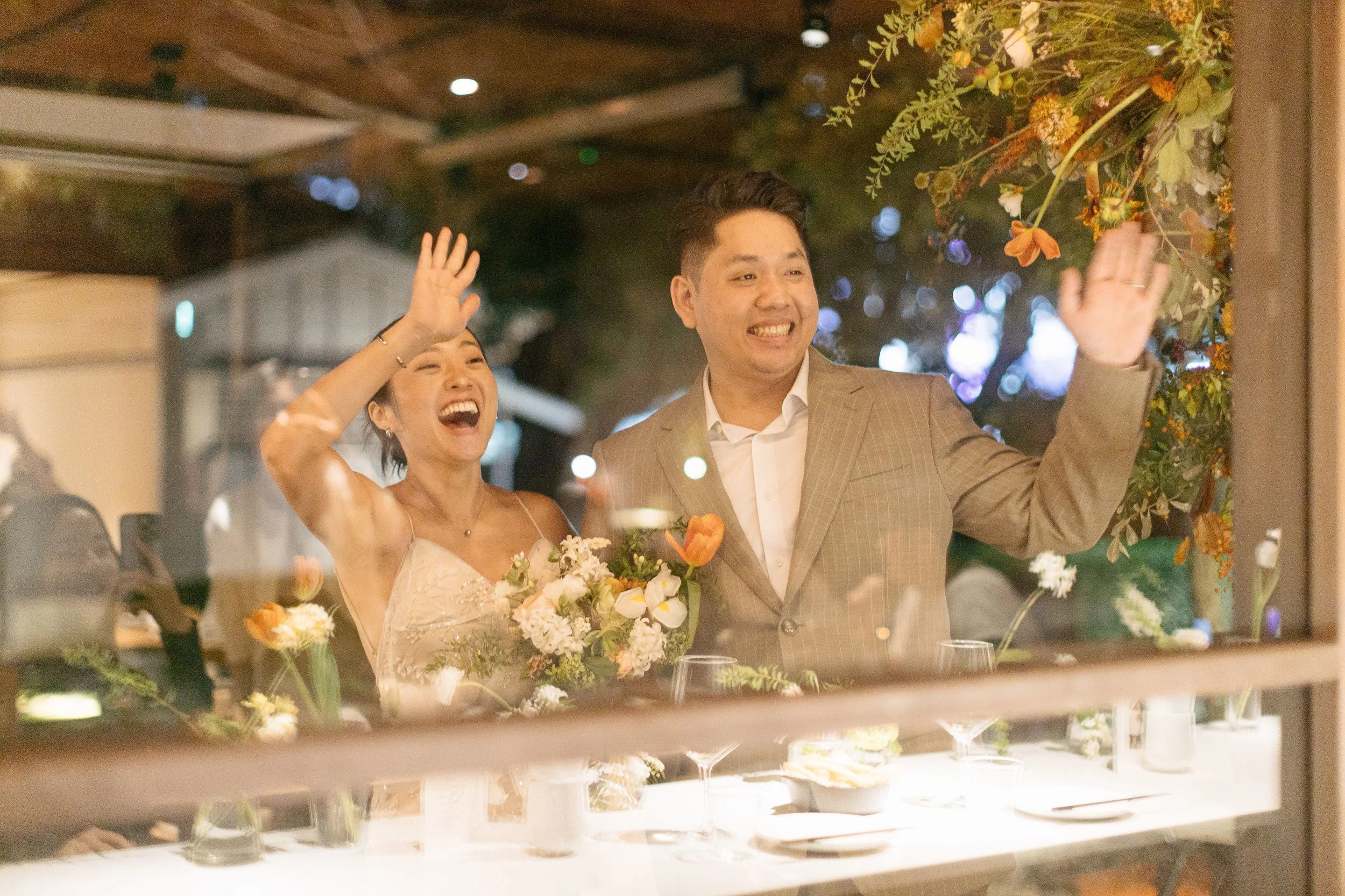 A joyful bride and groom waving and smiling at a wedding reception, decorated with flowers and warm lighting.