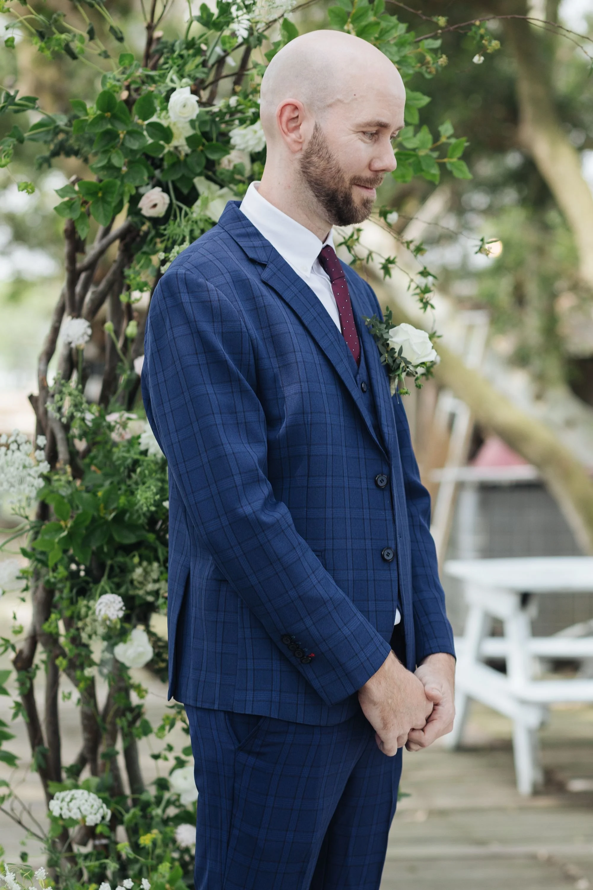 A man with a shaved head and beard is wearing a blue plaid suit with a white shirt and maroon tie. He is standing outdoors in front of an arch made of branches and white flowers, with a blurred background of trees and outdoor furniture.