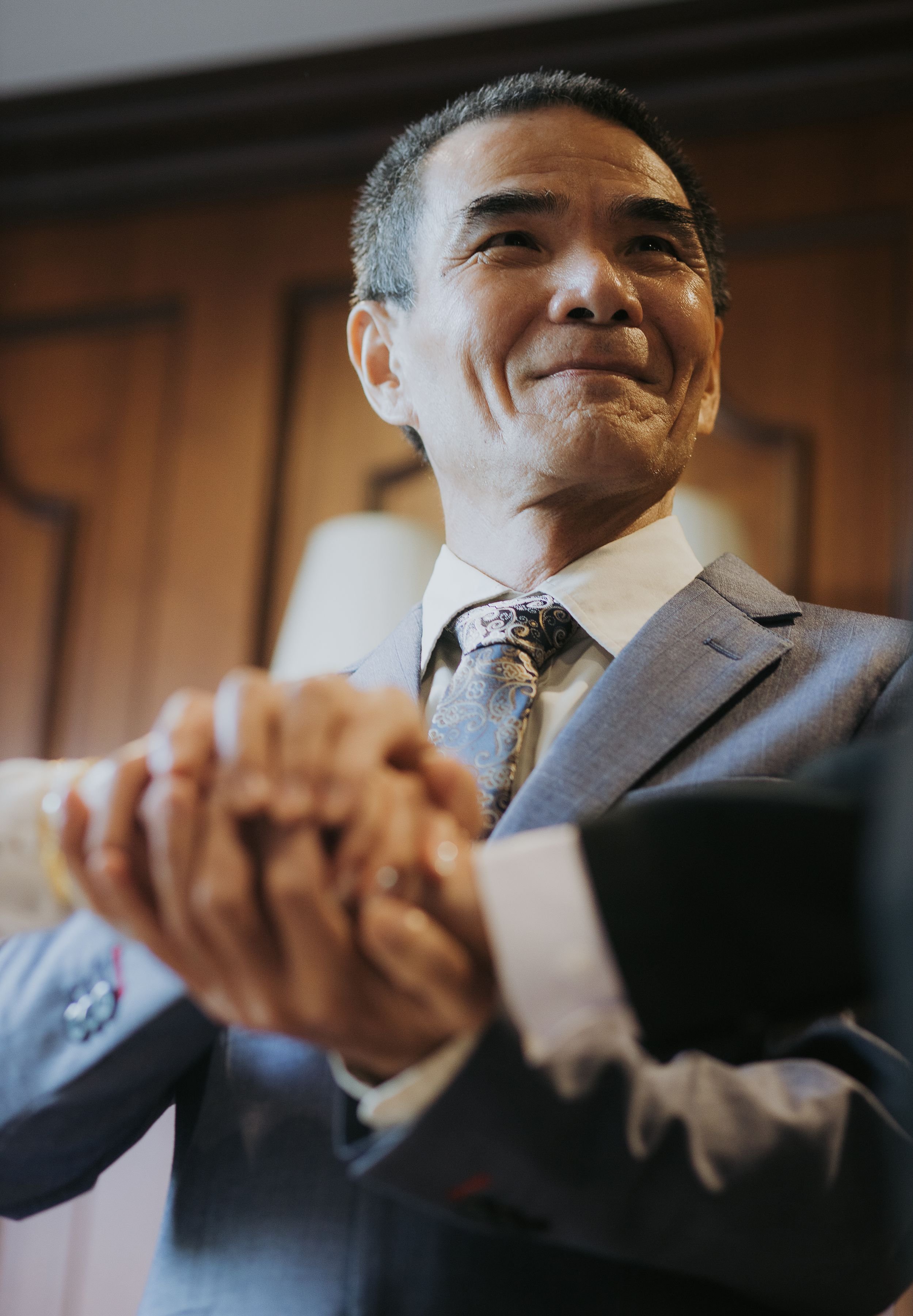 Close-up of a middle-aged Asian man in a suit, smiling while holding hands with a young girl in a gray sweater, in a formal setting with wood-paneled walls.