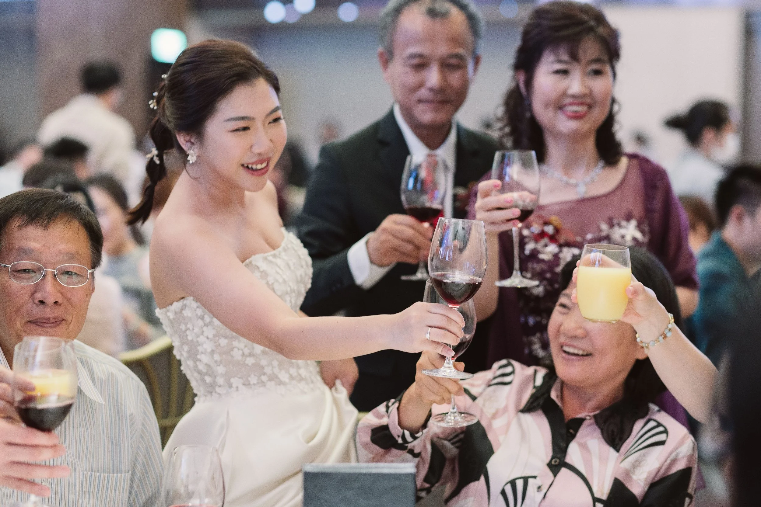 A group of people, including a bride in a white dress, are smiling and toasting with glasses of red wine, orange juice, and other drinks at a celebration or wedding reception.