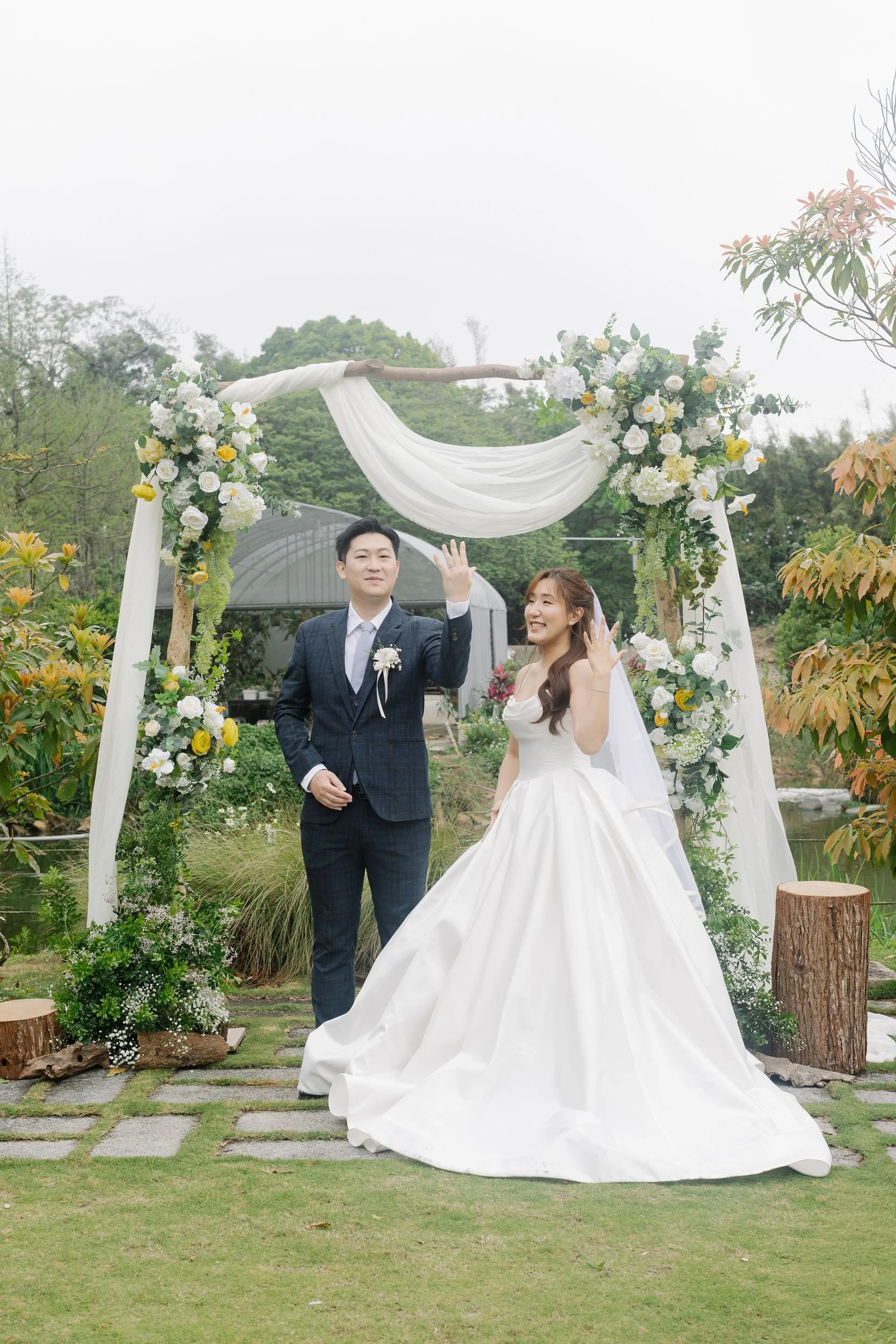 A bride and groom standing outdoors under a floral arch at their wedding, surrounded by greenery and trees.