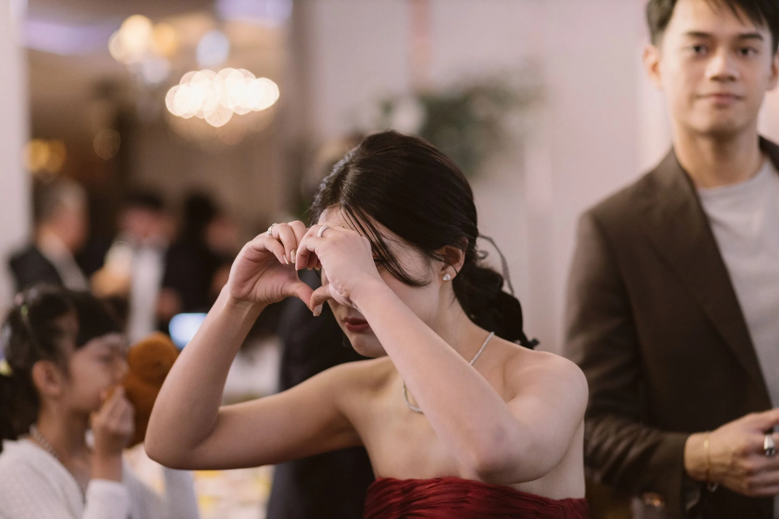 A woman with dark hair and pearl earrings making a heart shape with her hands, with a man and other people in the background at a social event.