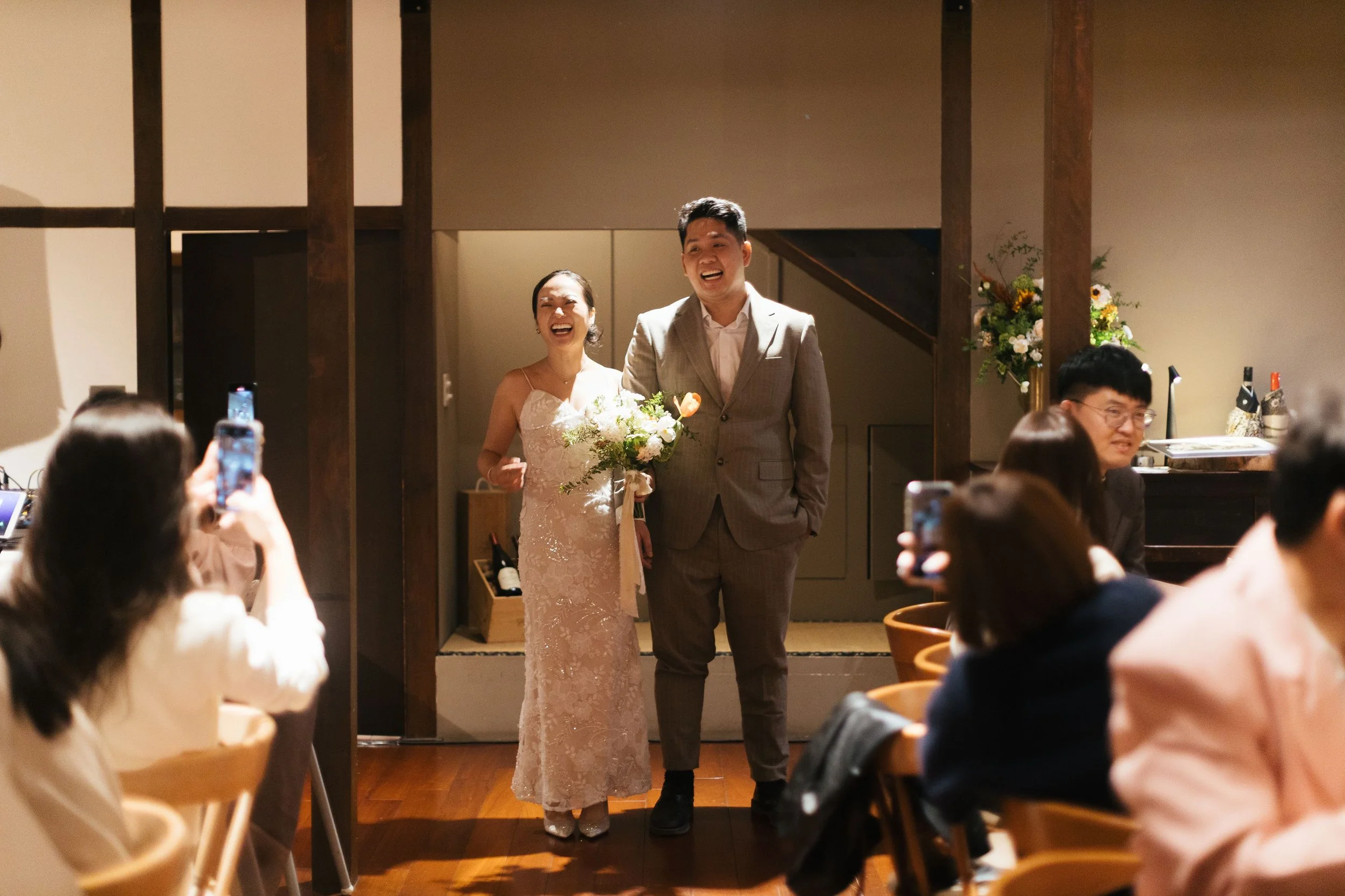 A bride and groom smiling as they walk into a wedding reception surrounded by seated guests taking photos, with a floral arrangement in the background.
