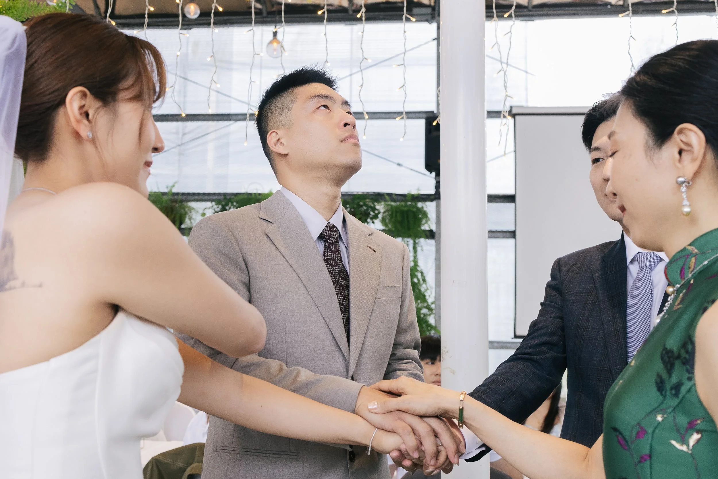 A group of professionally dressed individuals engaged in a handshake, with a focus on a woman in a white dress and a man in a beige suit exchanging handshakes in an indoor setting with hanging lights and windows in the background.