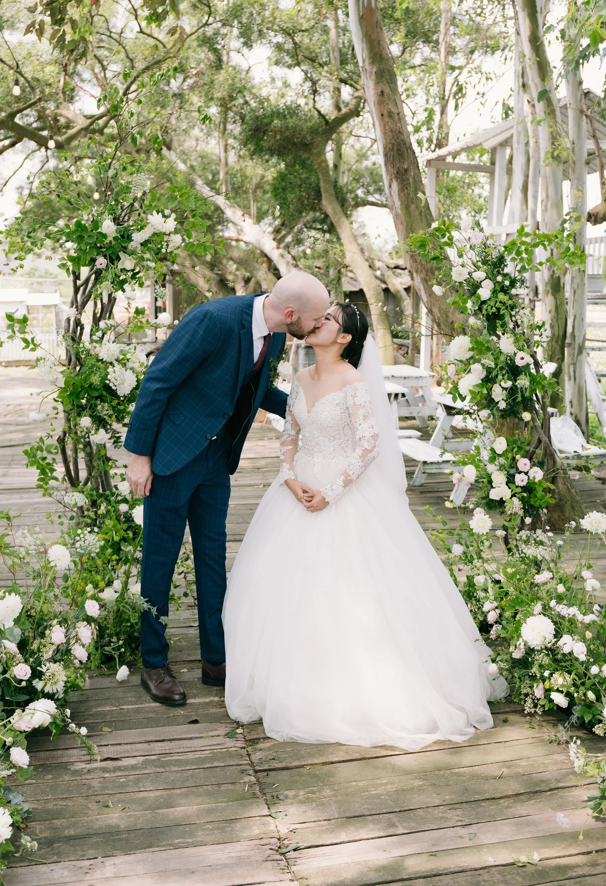 A newlywed couple sharing a kiss beneath a floral arch at an outdoor wedding. The groom is bald and wearing a blue plaid suit, while the bride has dark hair with a white veil and lace wedding dress. There are trees and greenery surrounding them, with