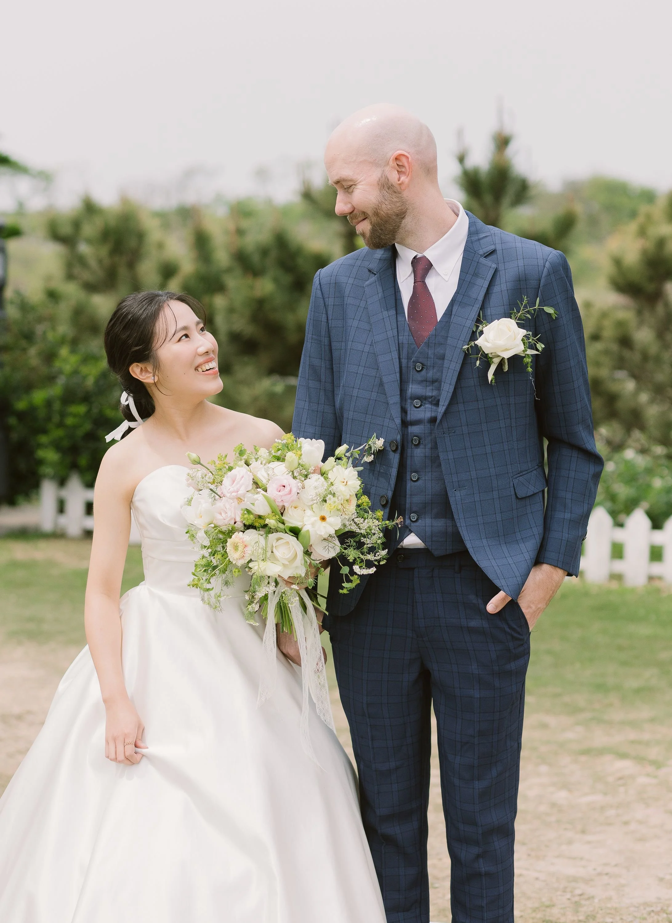 A bride and groom on their wedding day outdoors, with the bride holding a bouquet of white and pink flowers, and both looking at each other smiling.