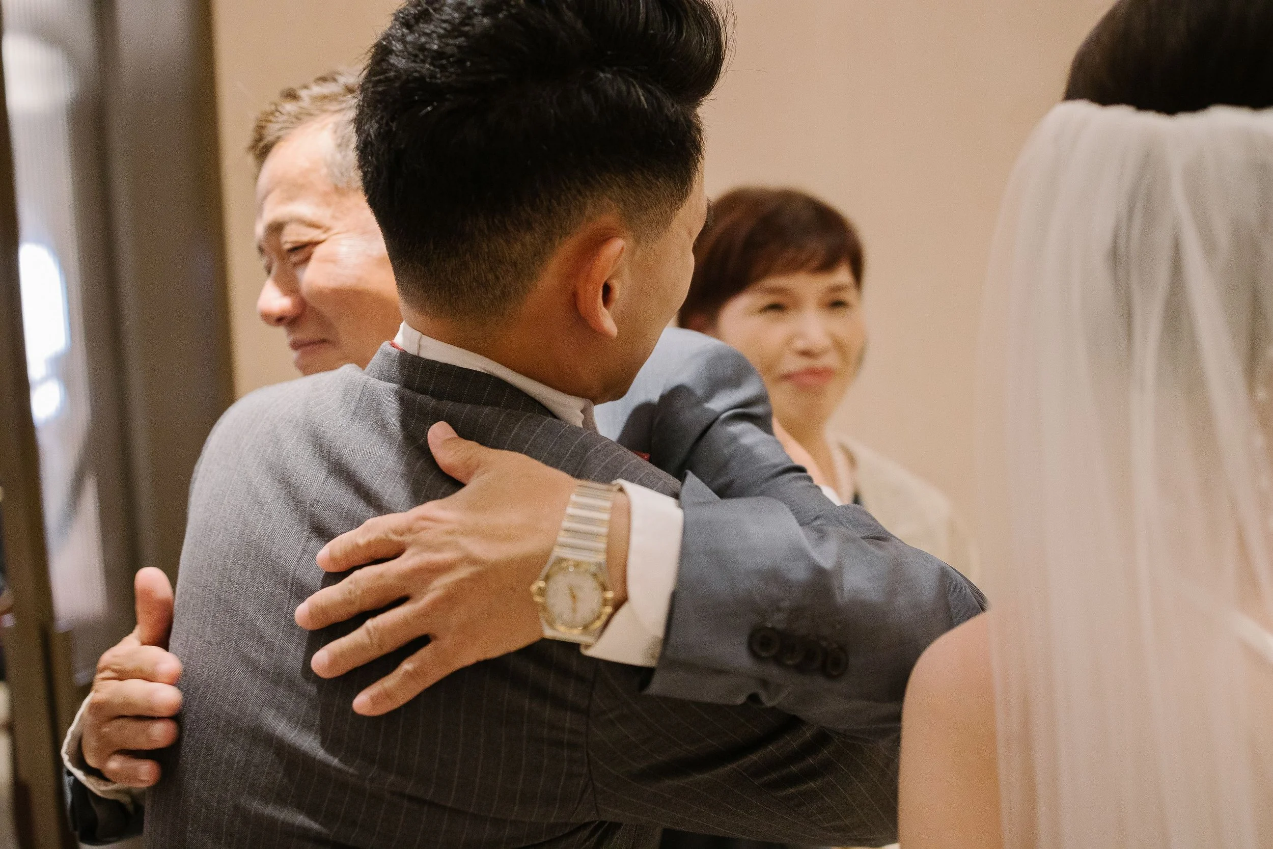 Two men in suits hugging each other, with one man wearing a gold watch, and a woman smiling in the background.