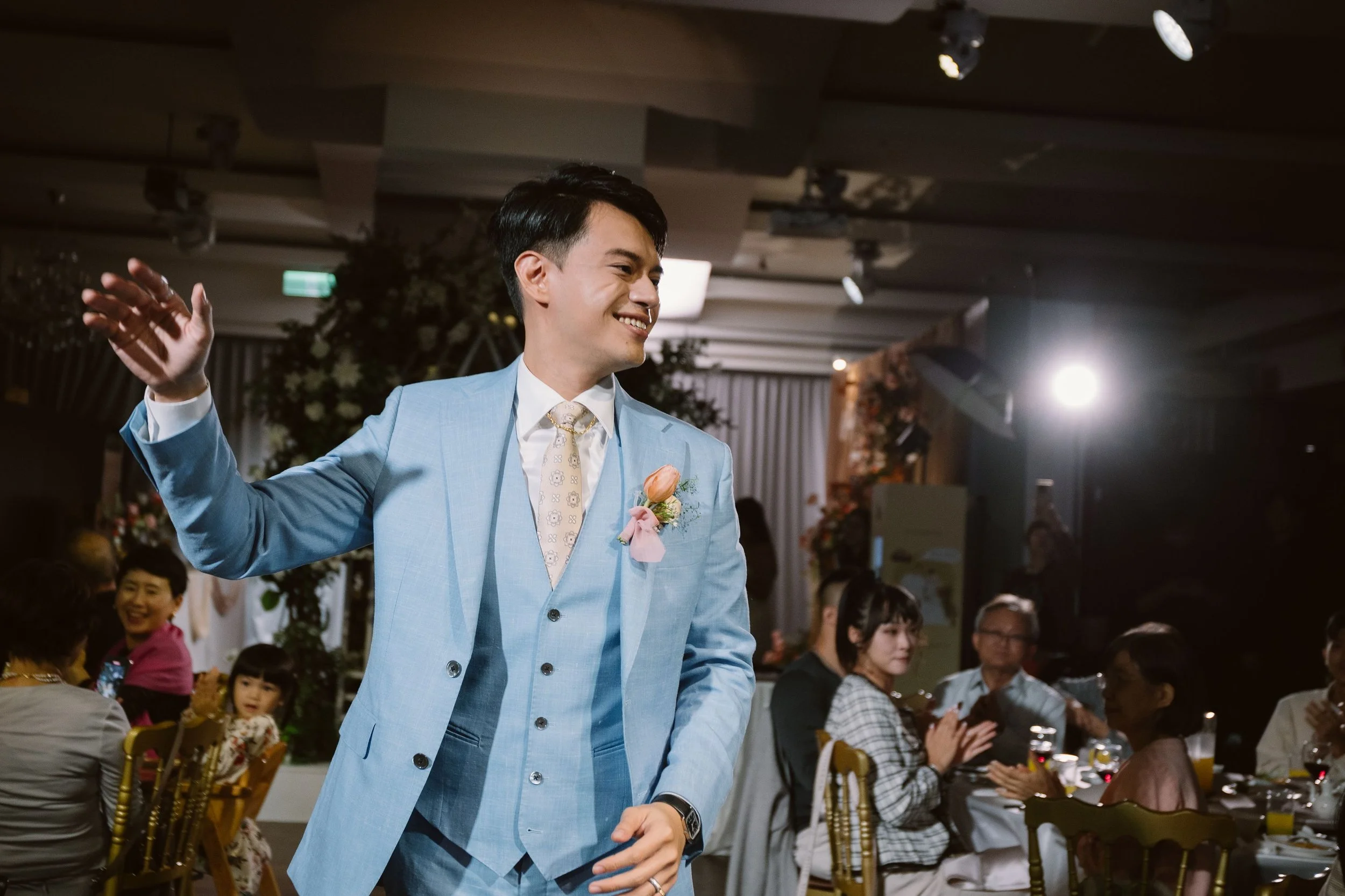A man in a light blue suit smiling and waving at a celebration or wedding event. People seated around tables indoors.