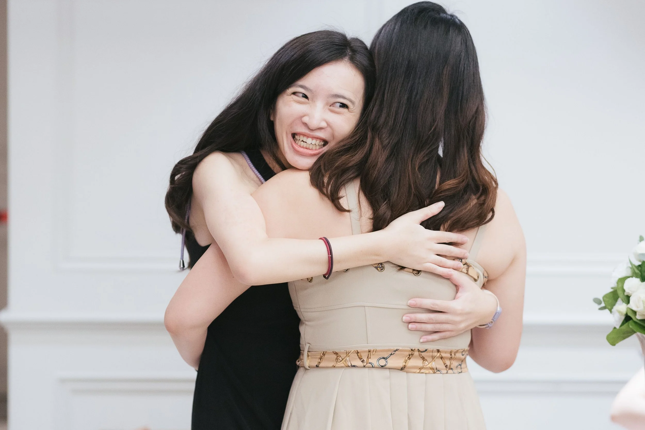 Two women hugging, one smiling and showing her teeth, the other with her back to the camera, in a bright indoor setting.