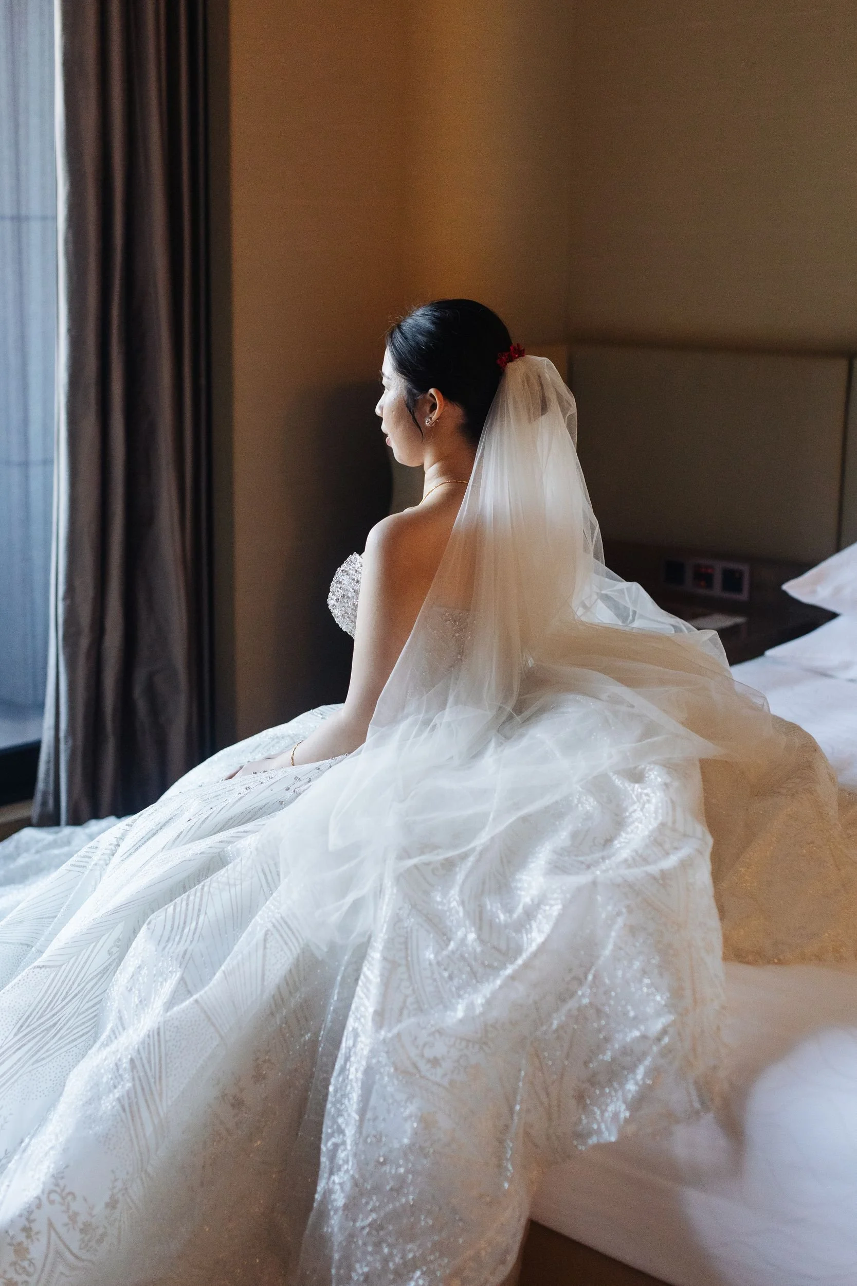A bride sitting on a bed, facing a window with closed curtains, wearing a wedding dress with a veil, in a dimly lit hotel room.