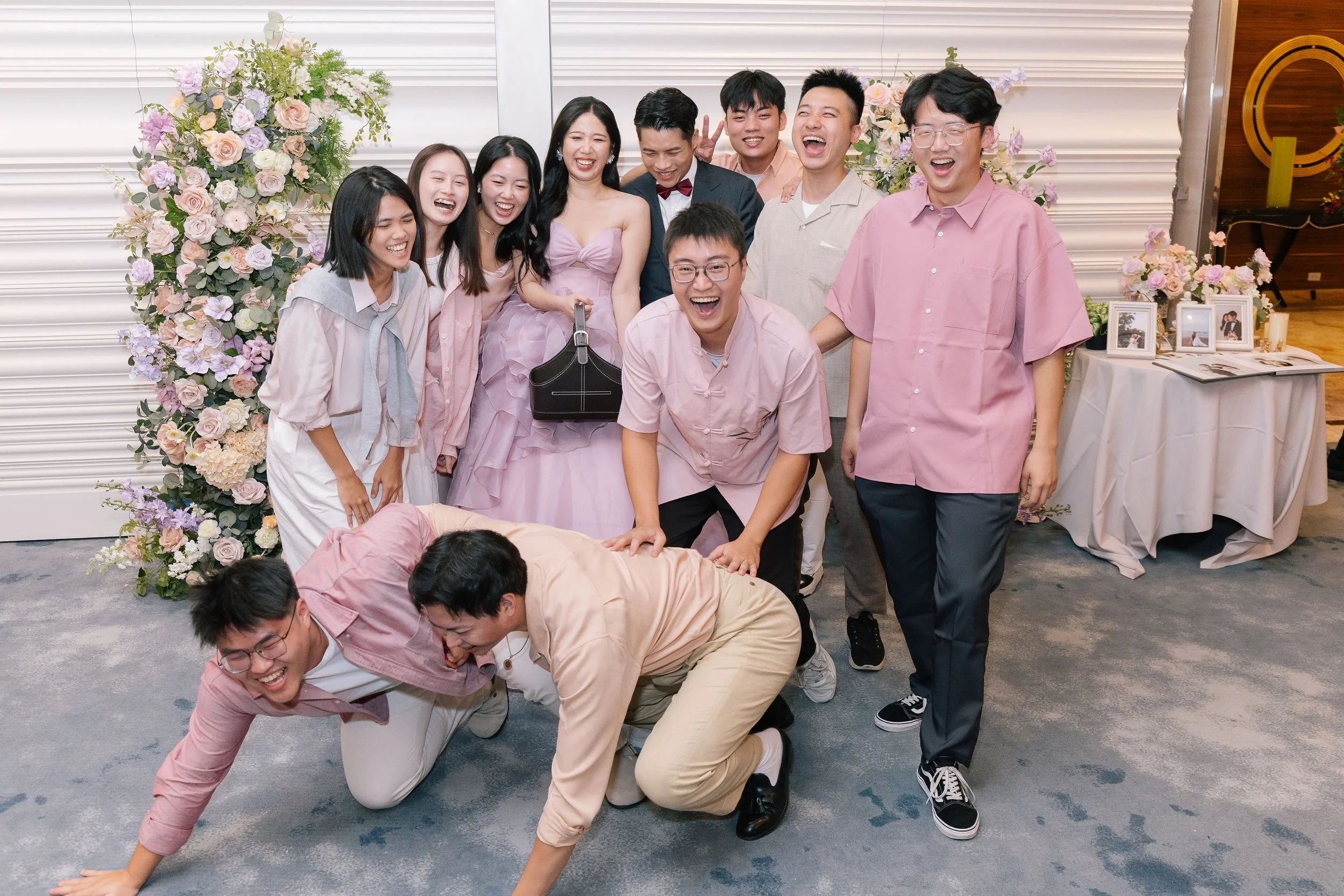 A group of people are posing for a photo at a celebration, with some standing and others on the floor doing a playful pose that resembles crawling or team-building activity. There are floral arrangements and framed photographs on a table in the backg