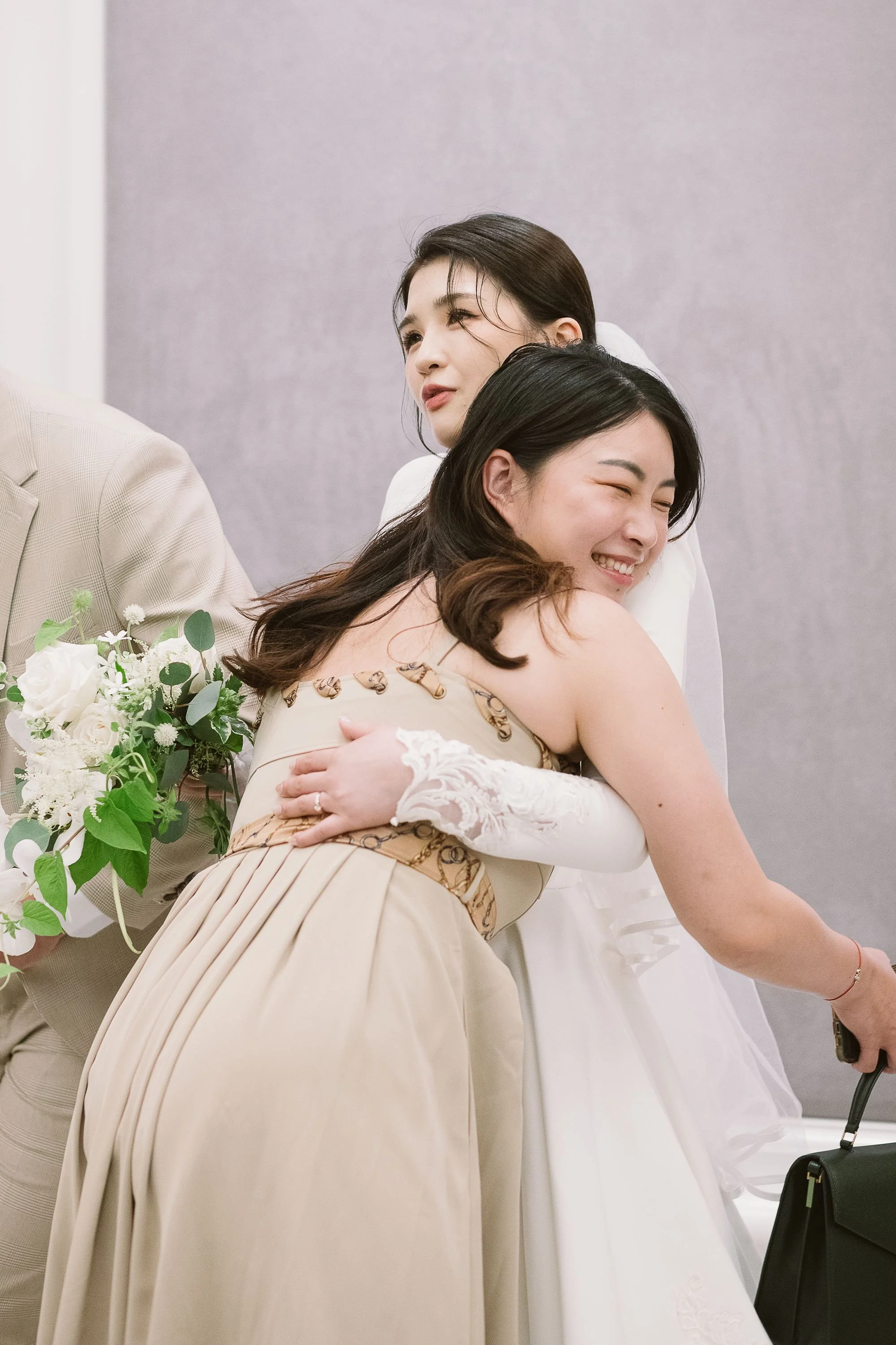 Two women hugging each other at a wedding or celebration, one wearing a beige dress and the other in a white dress, both smiling and embracing warmly.