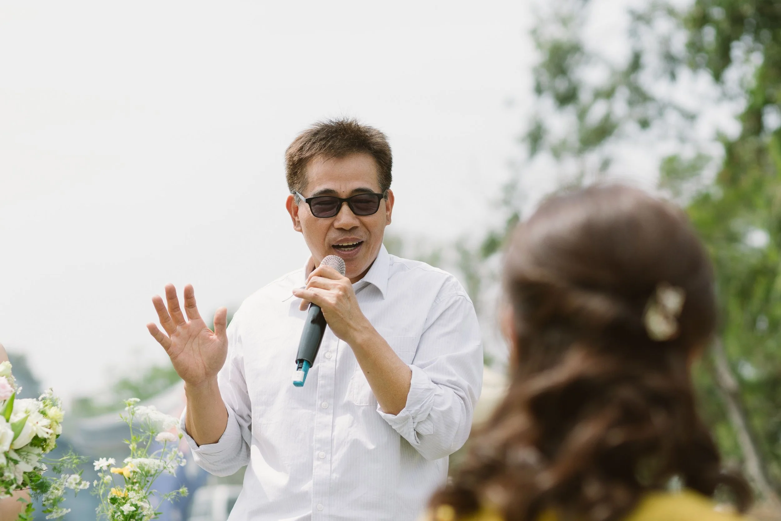 A man wearing sunglasses and a white shirt holding a microphone and speaking outdoors, engaging with an audience, with blurred greenery in the background.