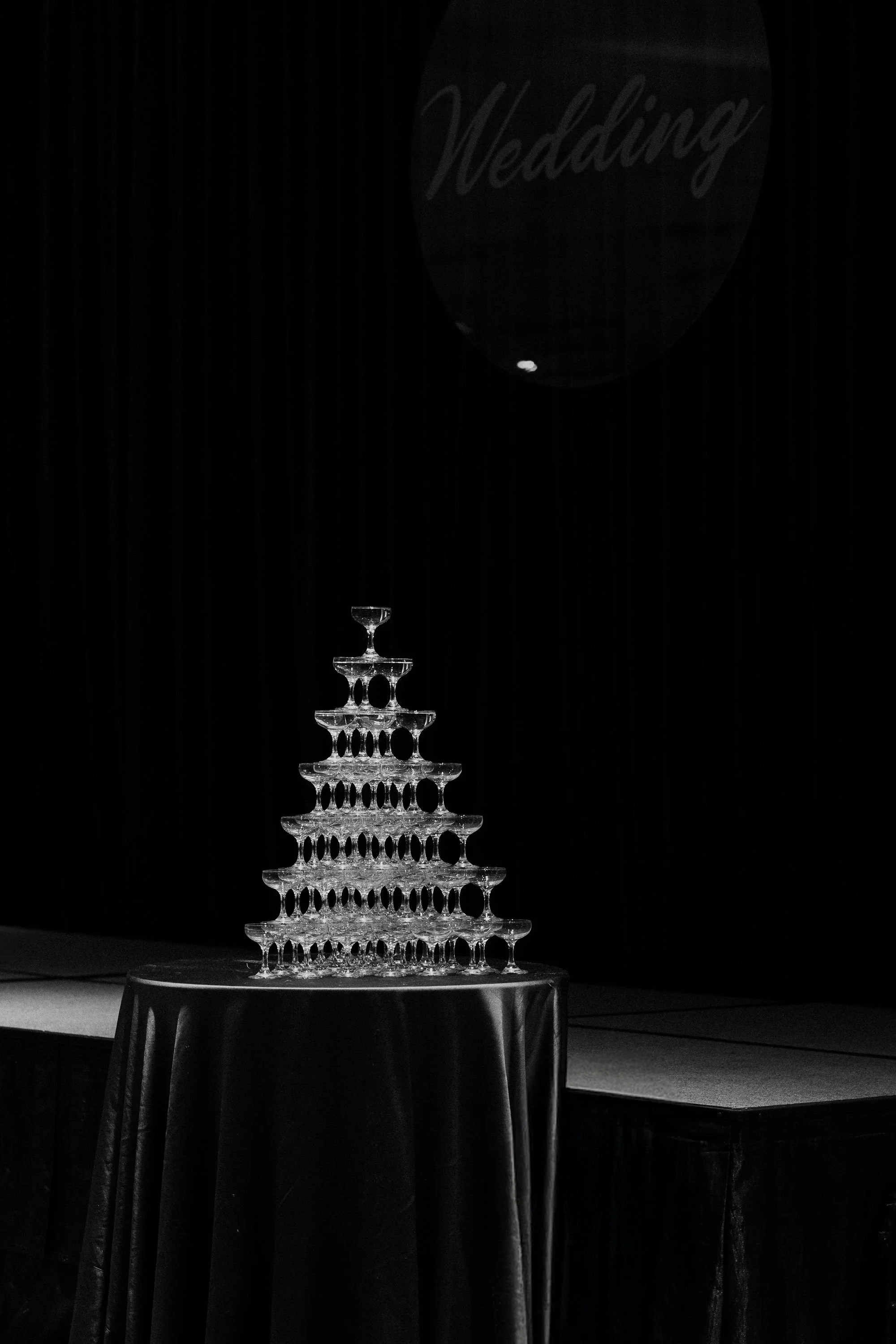 A multi-tiered crystal champagne tower on a table covered with a dark cloth, with a large black balloon above that has the word 'Wedding' written on it.