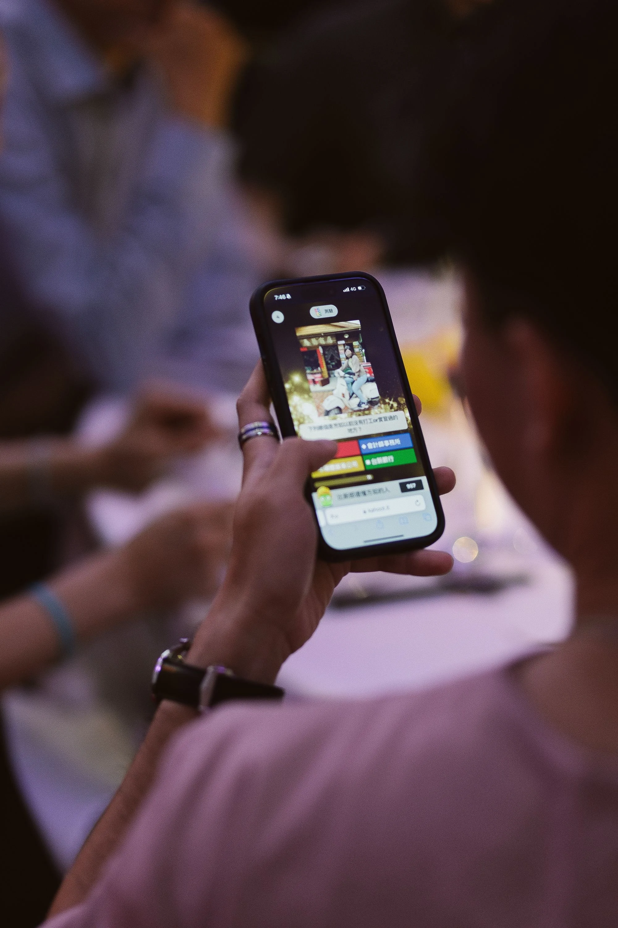 Close-up of a person's hand holding a smartphone, taking a picture of a scene that appears to be an outdoor or public setting with people and lights in the background.