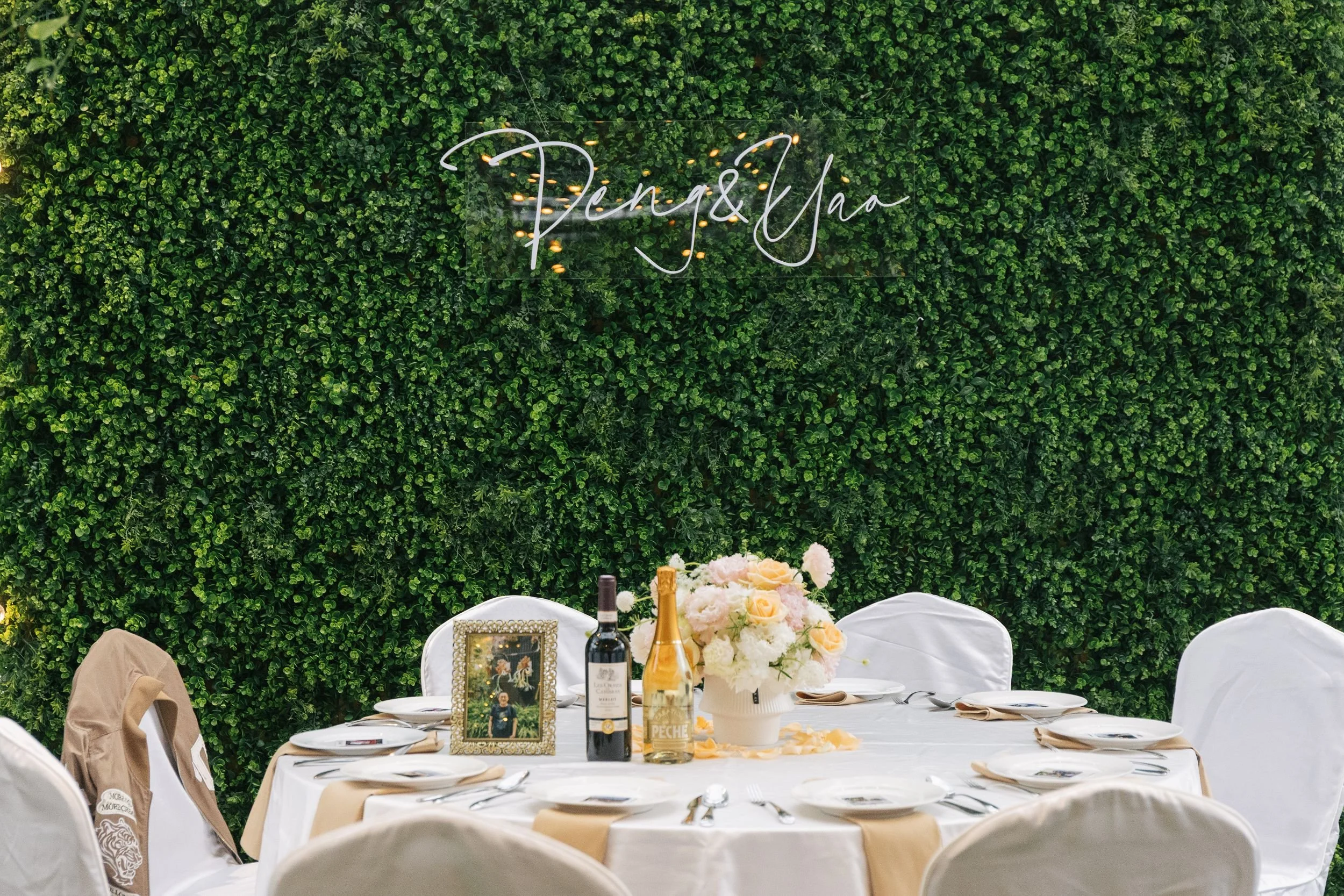 Wedding reception table with white tablecloths, floral centerpieces, framed photograph, wine bottles, and decorated chairs against a green leafy wall with a neon sign reading 'Penang & Jaa'.