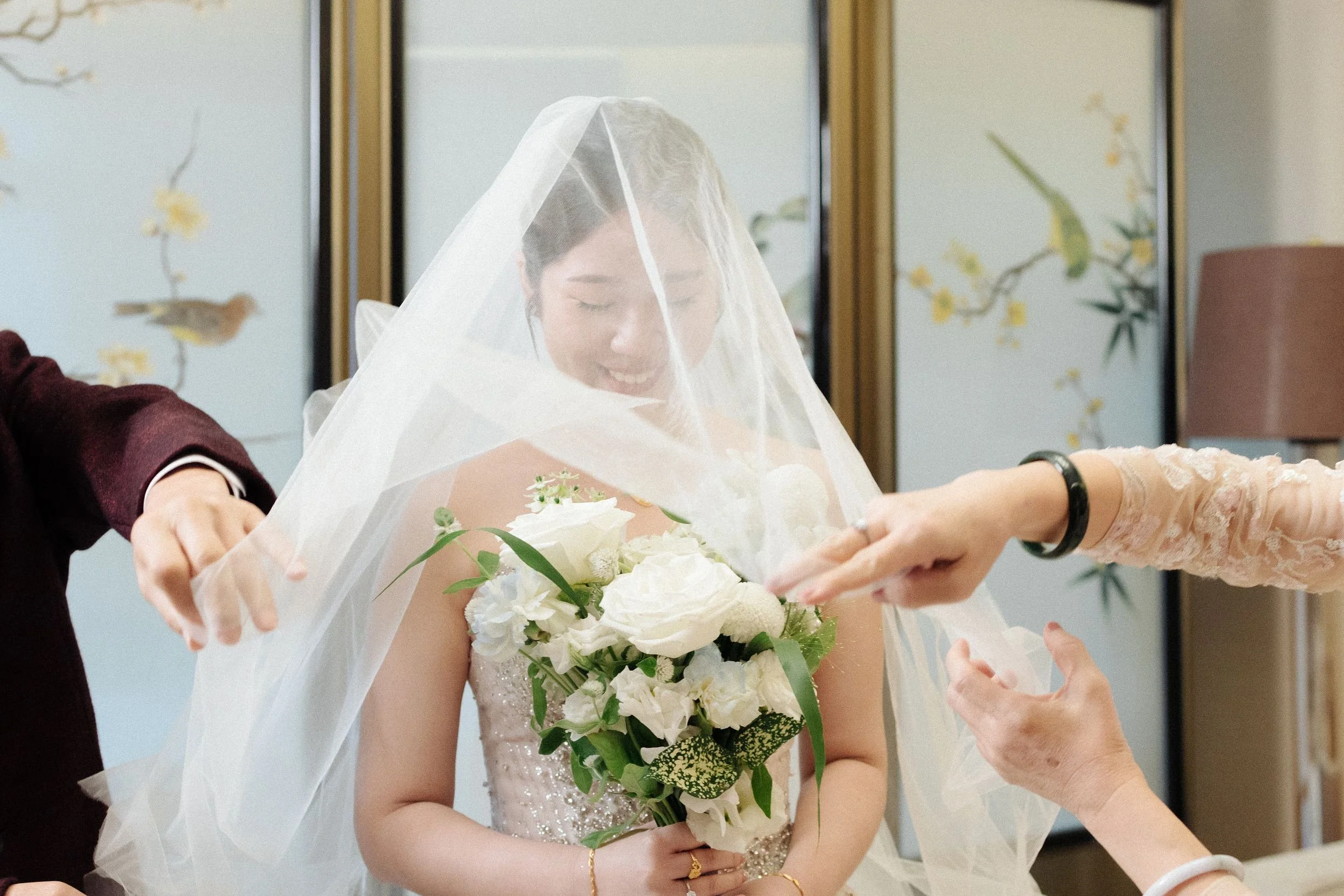 A smiling bride in a wedding dress holding a bouquet of white flowers, with people assisting her in placing a veil on her head.