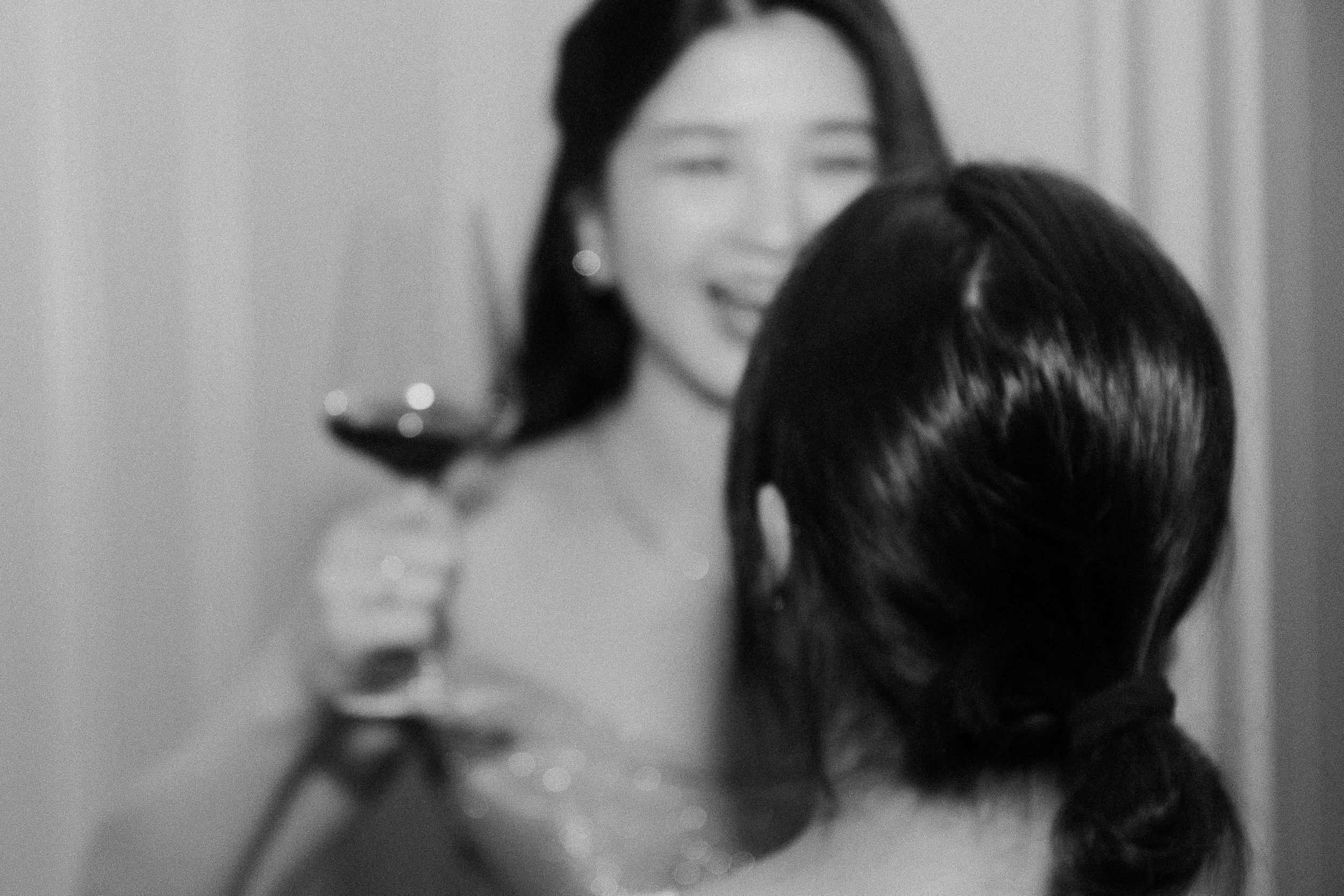 A woman smiling as her hair is being styled by another person in a salon, with soft focus and black-and-white filter.