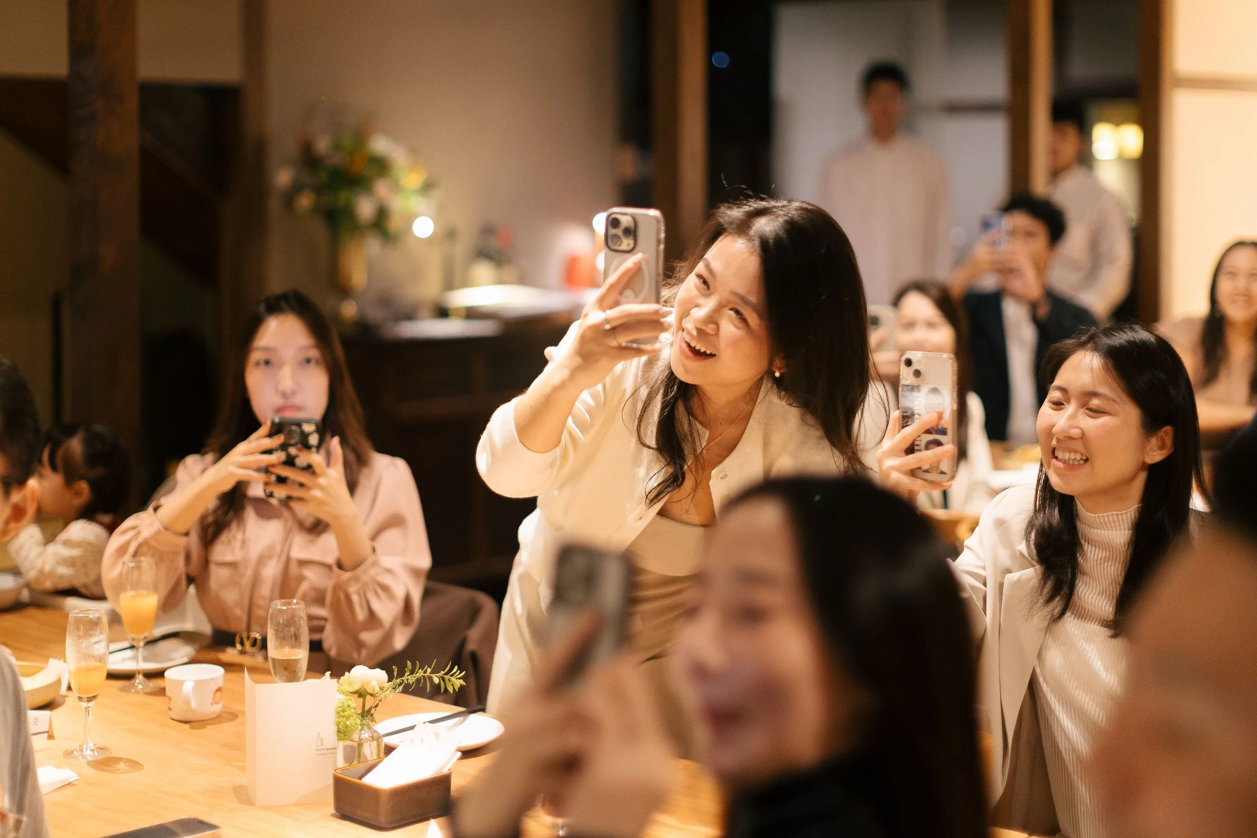 A group of women and men gathered around a dining table at a celebration, taking photos and smiling. The table has drinks, flowers, and place settings, and the atmosphere is warm and festive.