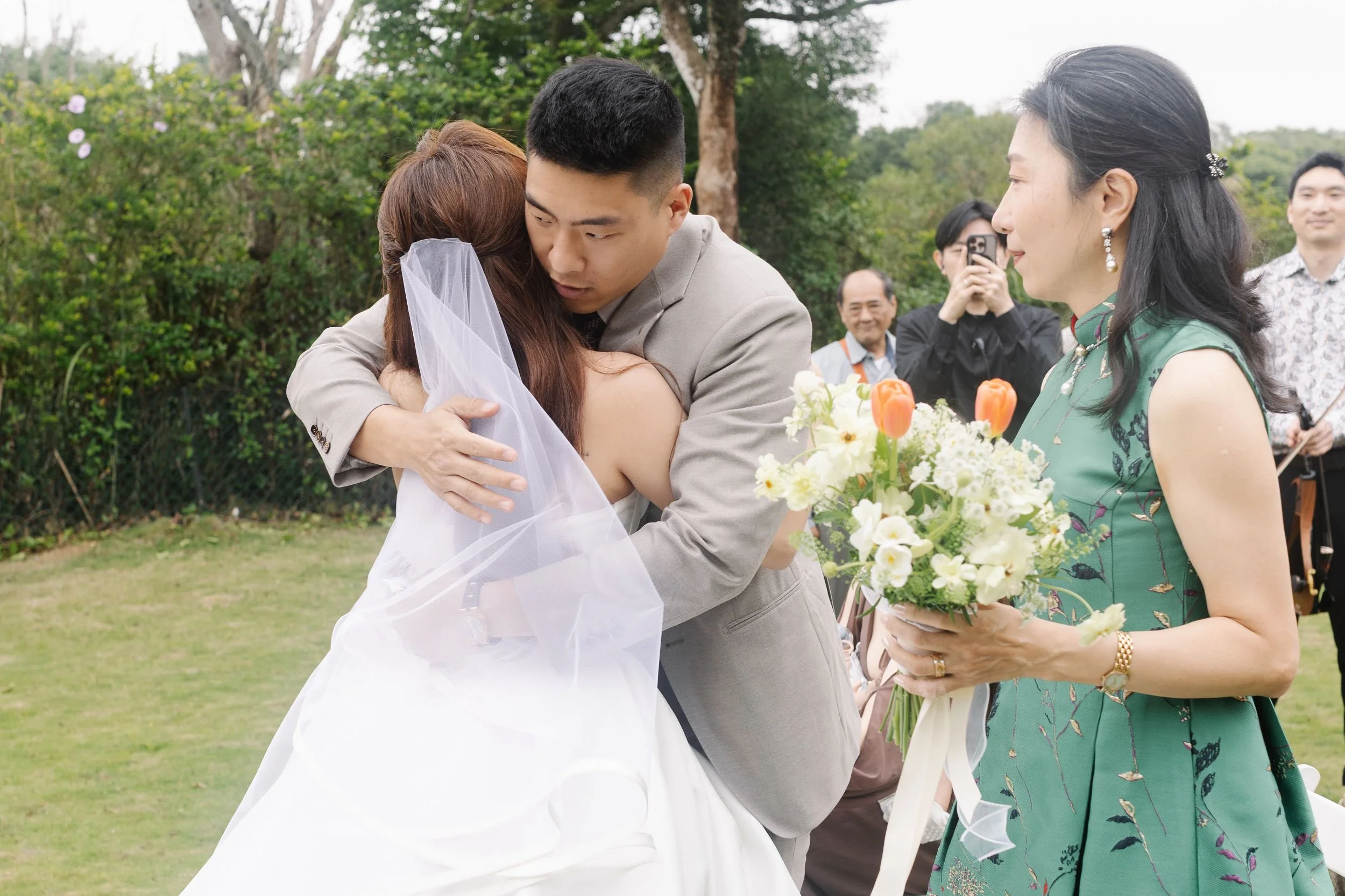 A groom and bride embrace during a wedding ceremony outdoors, surrounded by family and friends, with a woman holding a bouquet of flowers nearby.
