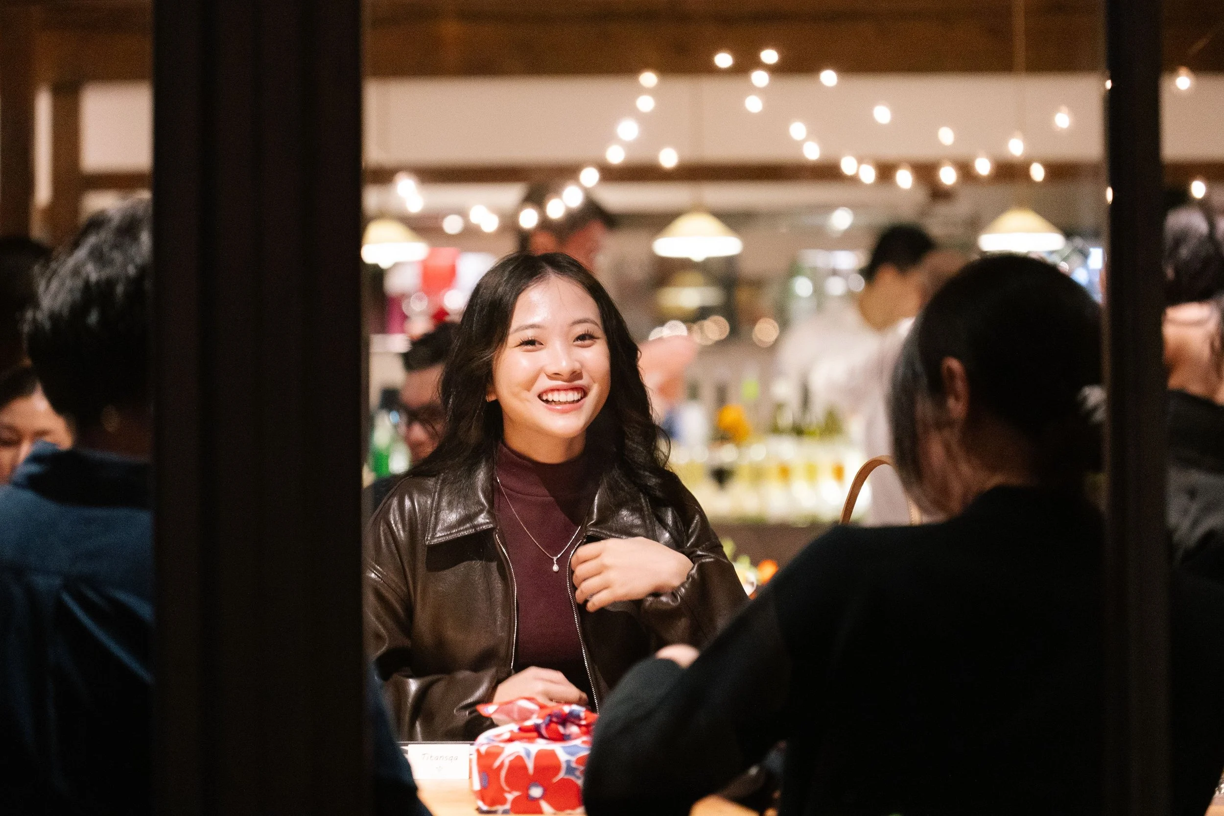 A young woman with long dark hair smiling at a social gathering, wearing a leather jacket in a warmly lit room with other people in the background.