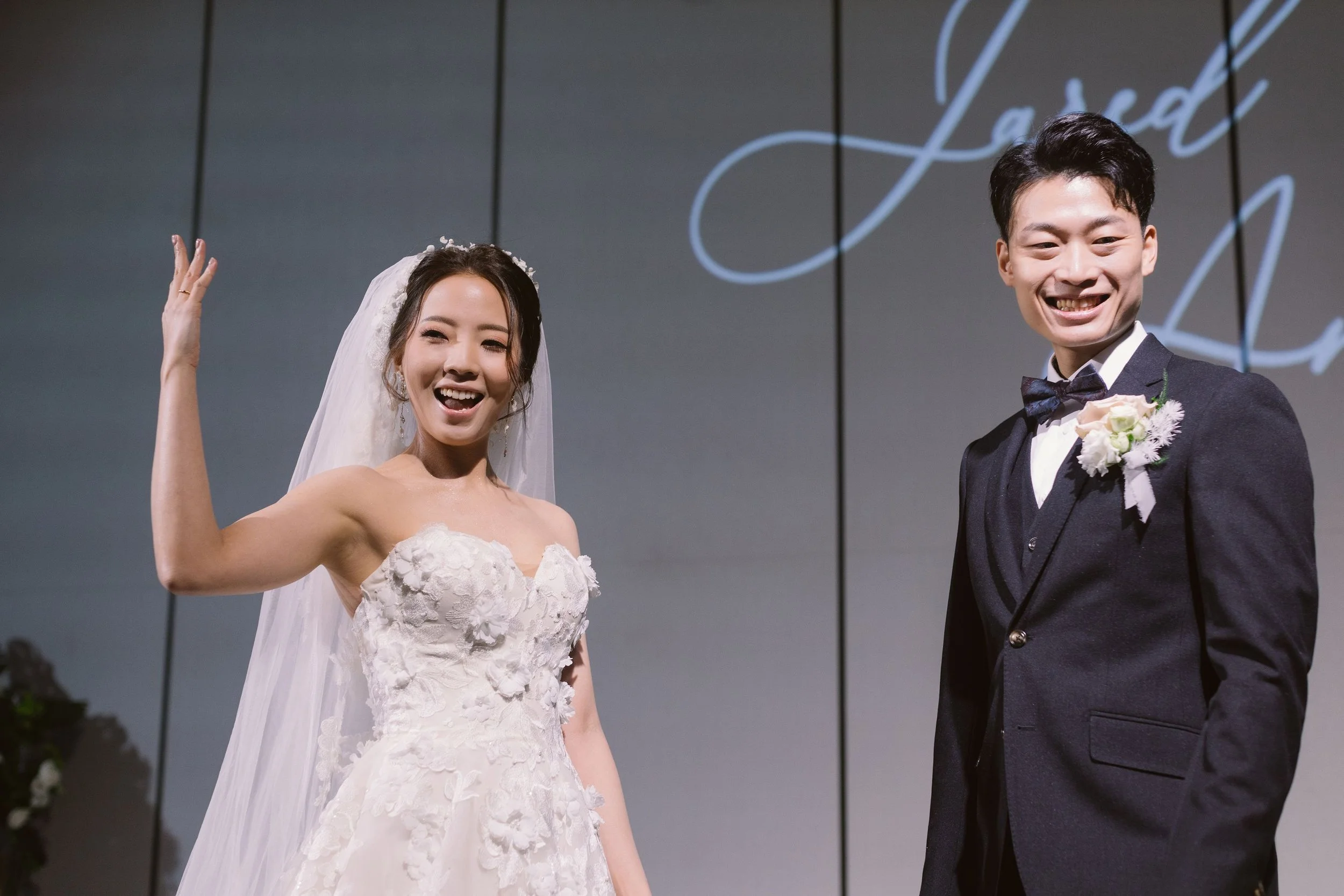 A bride and groom smiling at a wedding ceremony, with the bride in a strapless lace wedding gown and the groom in a black tuxedo with a bow tie, standing in front of a dark background with illuminated cursive writing.