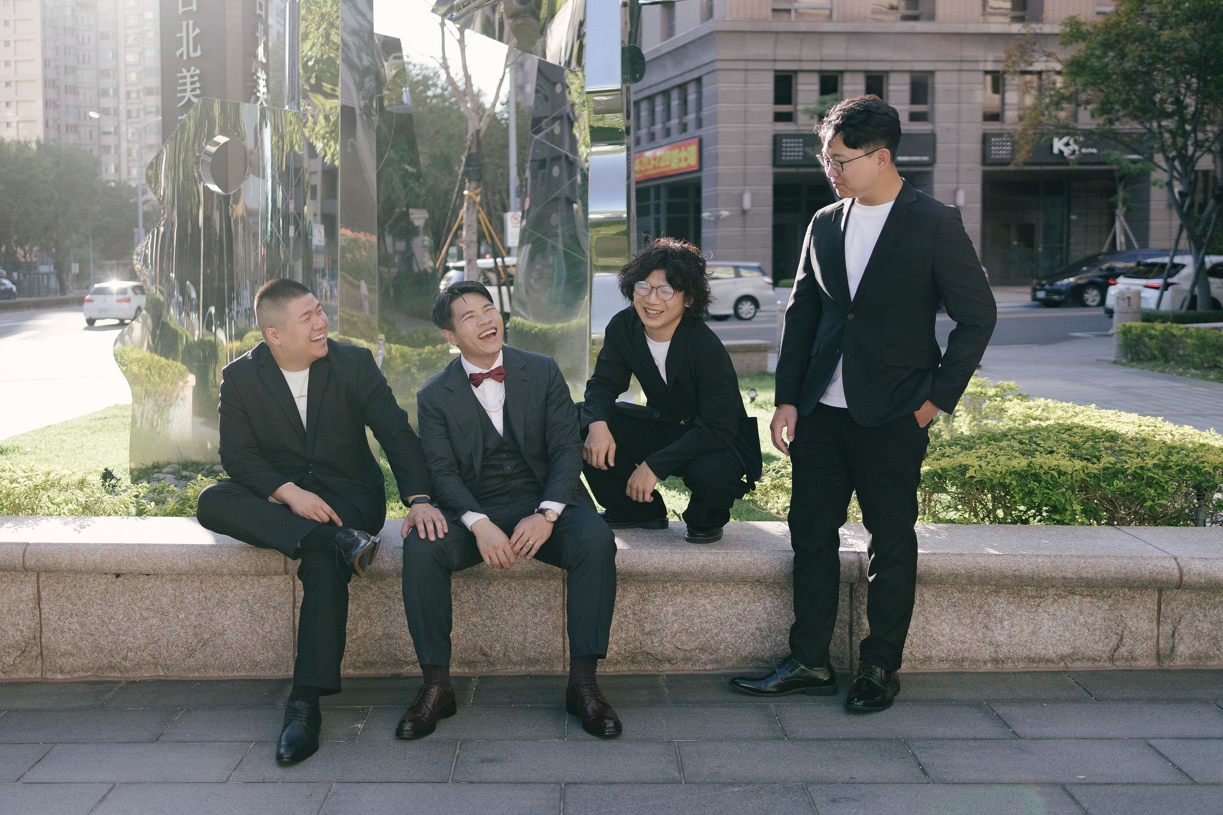 Five young men in formal and casual attire are gathered on a city sidewalk, smiling and laughing. Three are seated on a low wall, and two are standing. They are in front of a reflective metal sculpture with modern buildings and trees in the backgroun