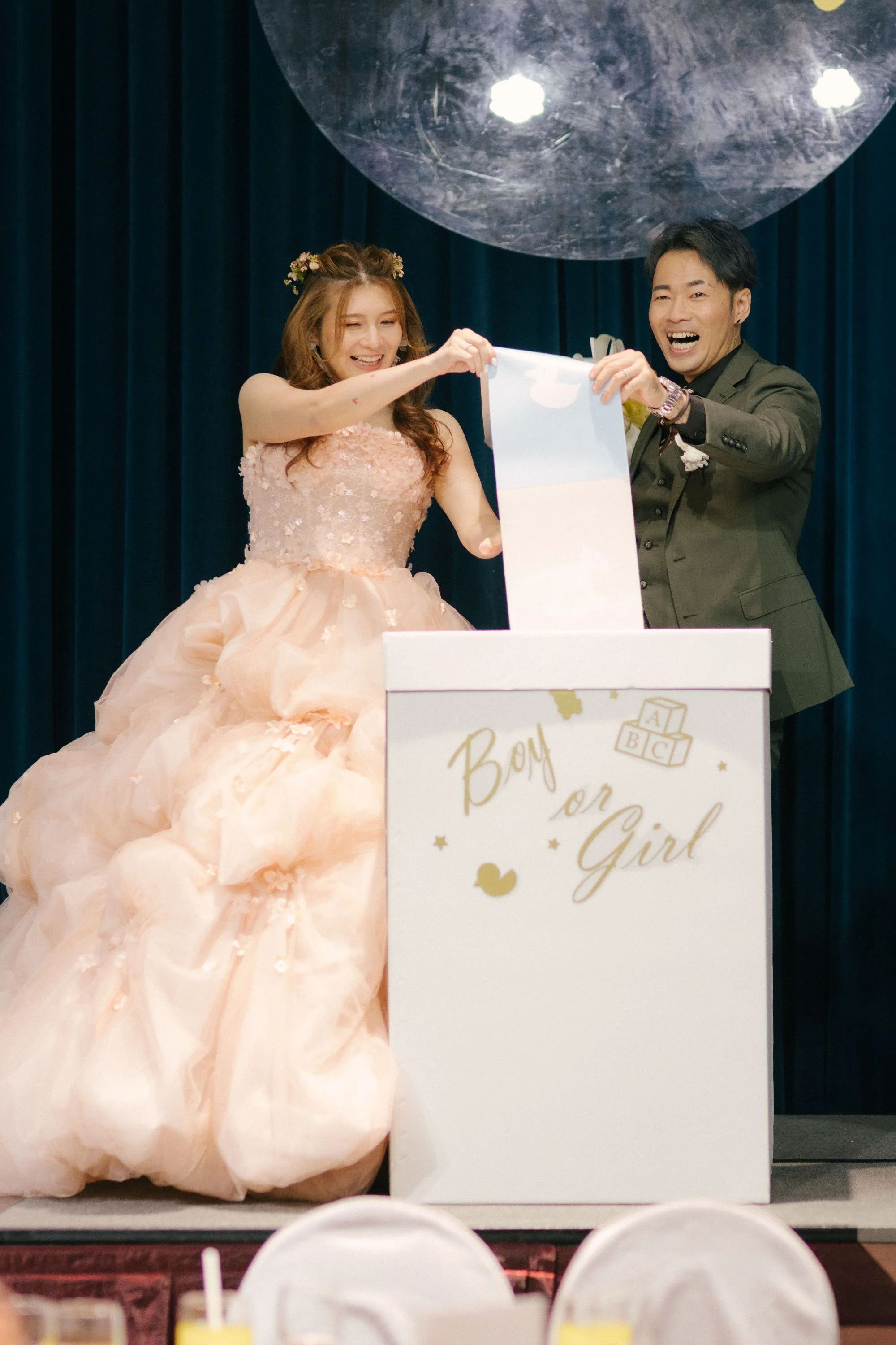 A woman in a pink ball gown and a man in a suit are smiling and holding a poster in front of a stage with a sign that says 'Boy or Girl'.