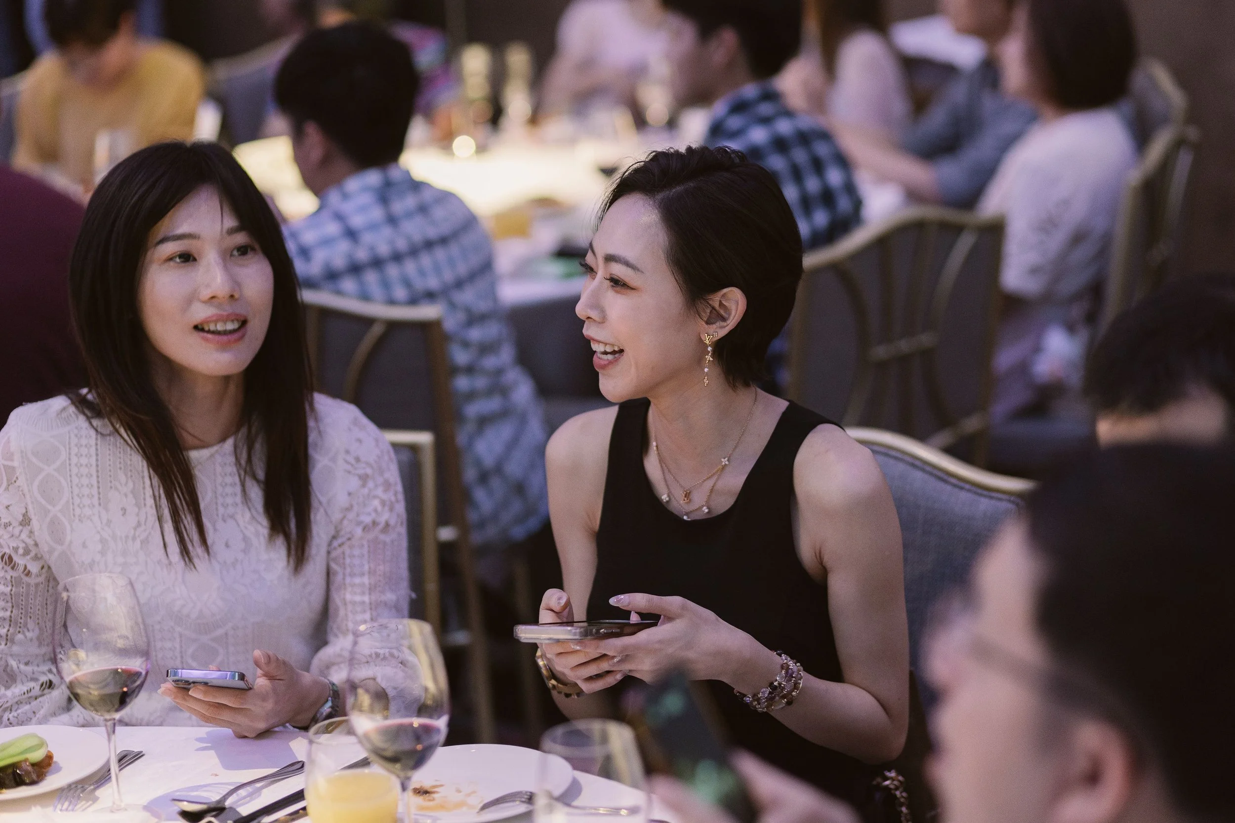 People socializing at a dinner party, with women engaging in conversation and using their phones, set in a well-lit indoor setting with a table of food and drinks.