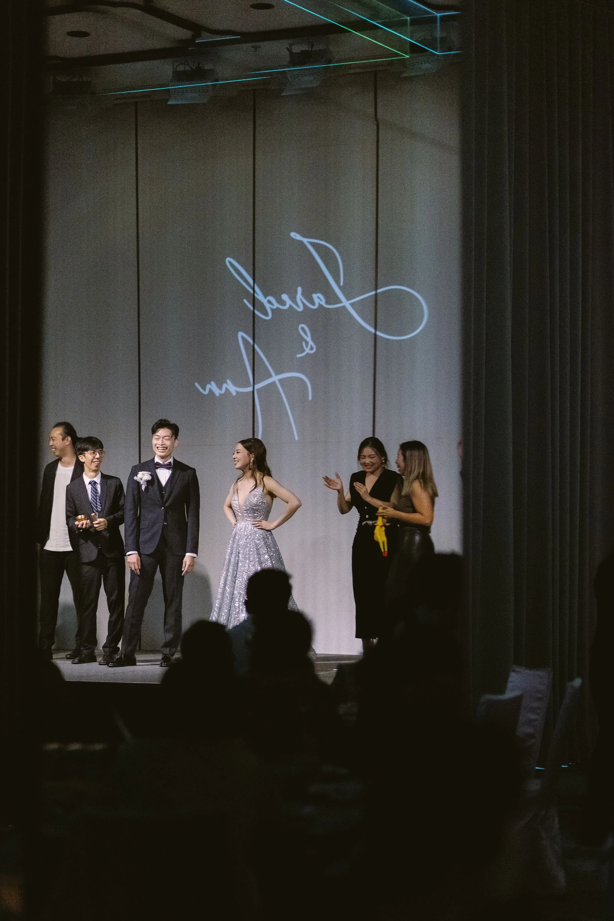 A group of people on stage at an event, including a man in a tuxedo, a woman in a silver gown, and others clapping and smiling in front of a large screen with cursive writing.