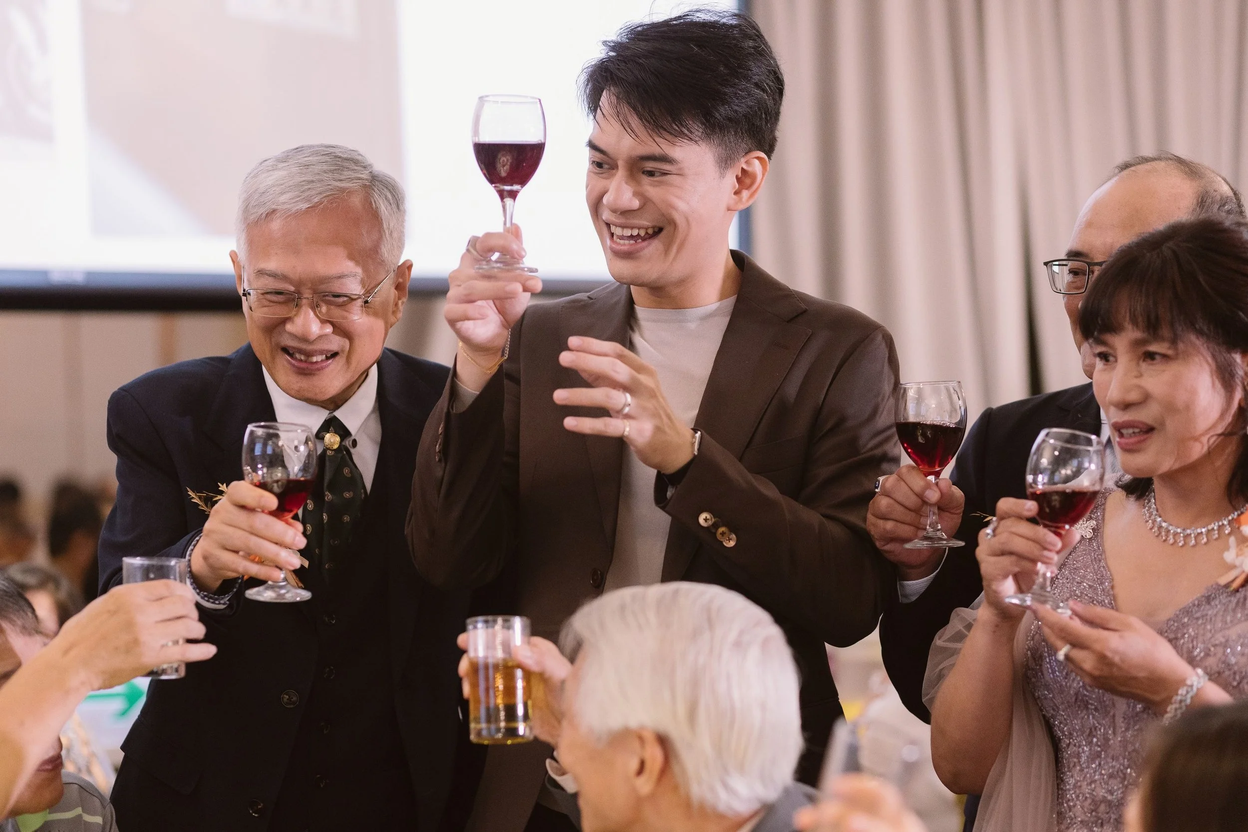 A group of people at a social gathering or celebration, holding glasses of red wine, with one man smiling and holding his glass, others smiling and engaging in conversation.
