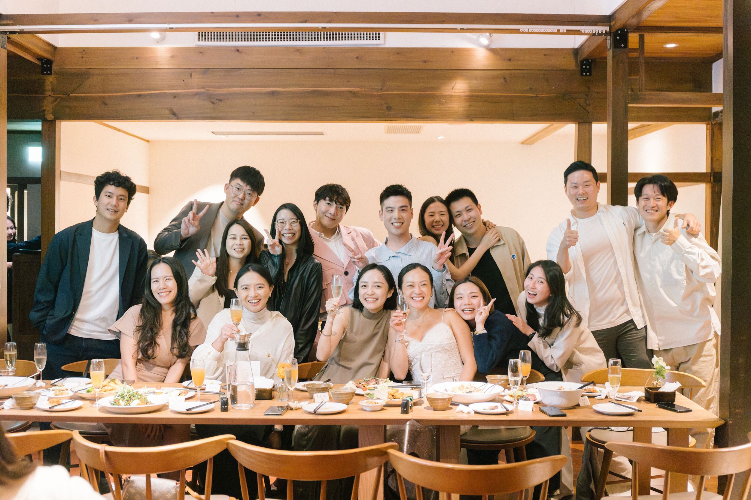 Group of people celebrating at a dinner party in a restaurant, smiling and making peace signs or thumbs-up gestures, with food and drinks on the table.