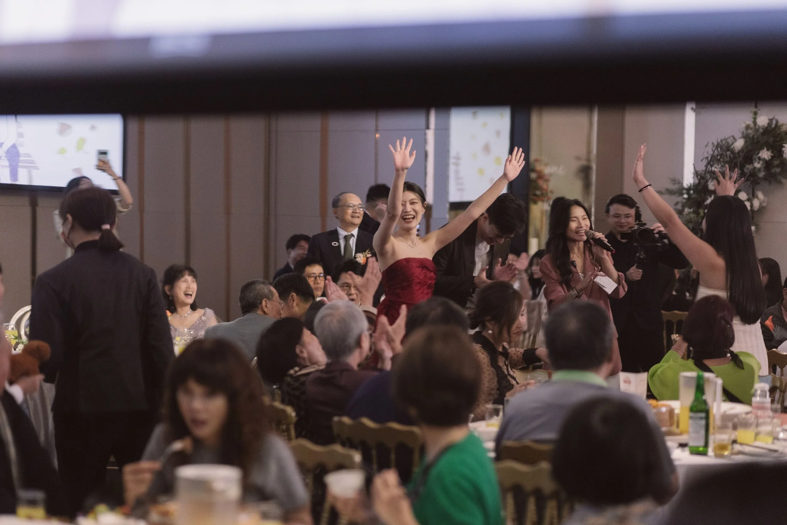 A celebration scene with a woman in a red dress raising her hands, surrounded by smiling guests at an indoor event.