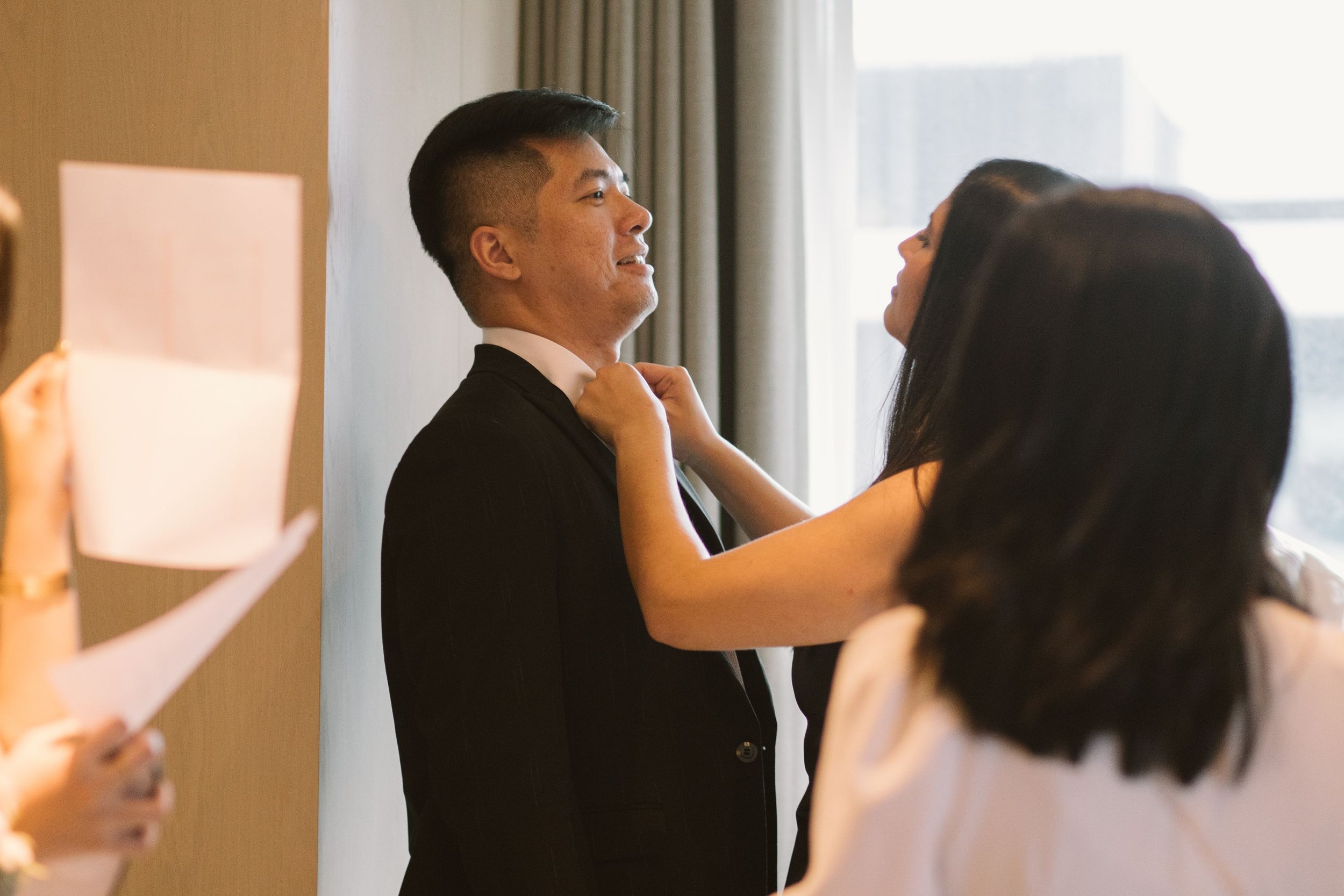 A man in a suit is adjusting his tie while a woman helps him, standing near a window with curtains.