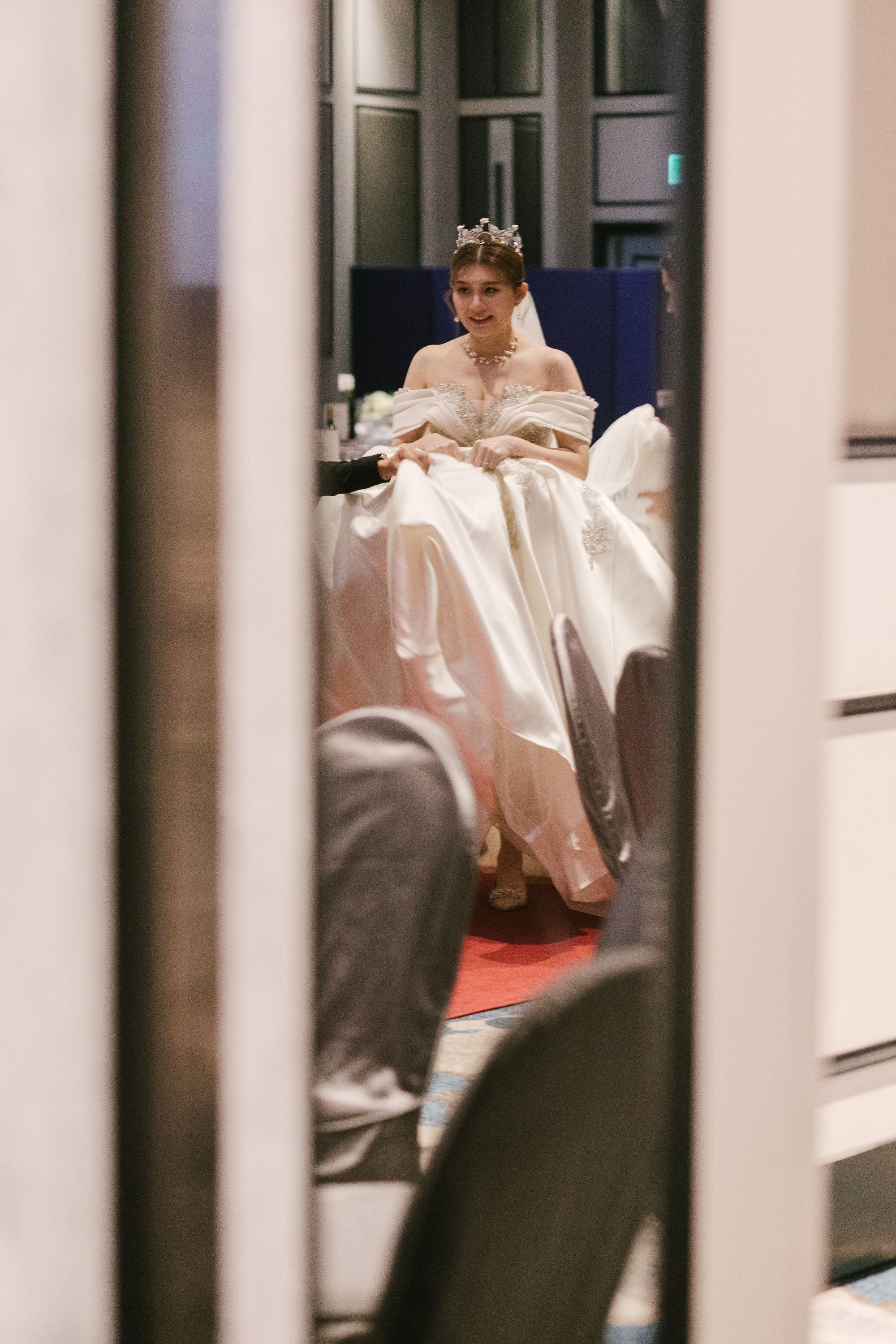 A bride wearing a wedding gown, pearl necklace, and tiara, sitting and smiling, as seen through a mirror reflection in a room with dark blue walls and gray chairs.