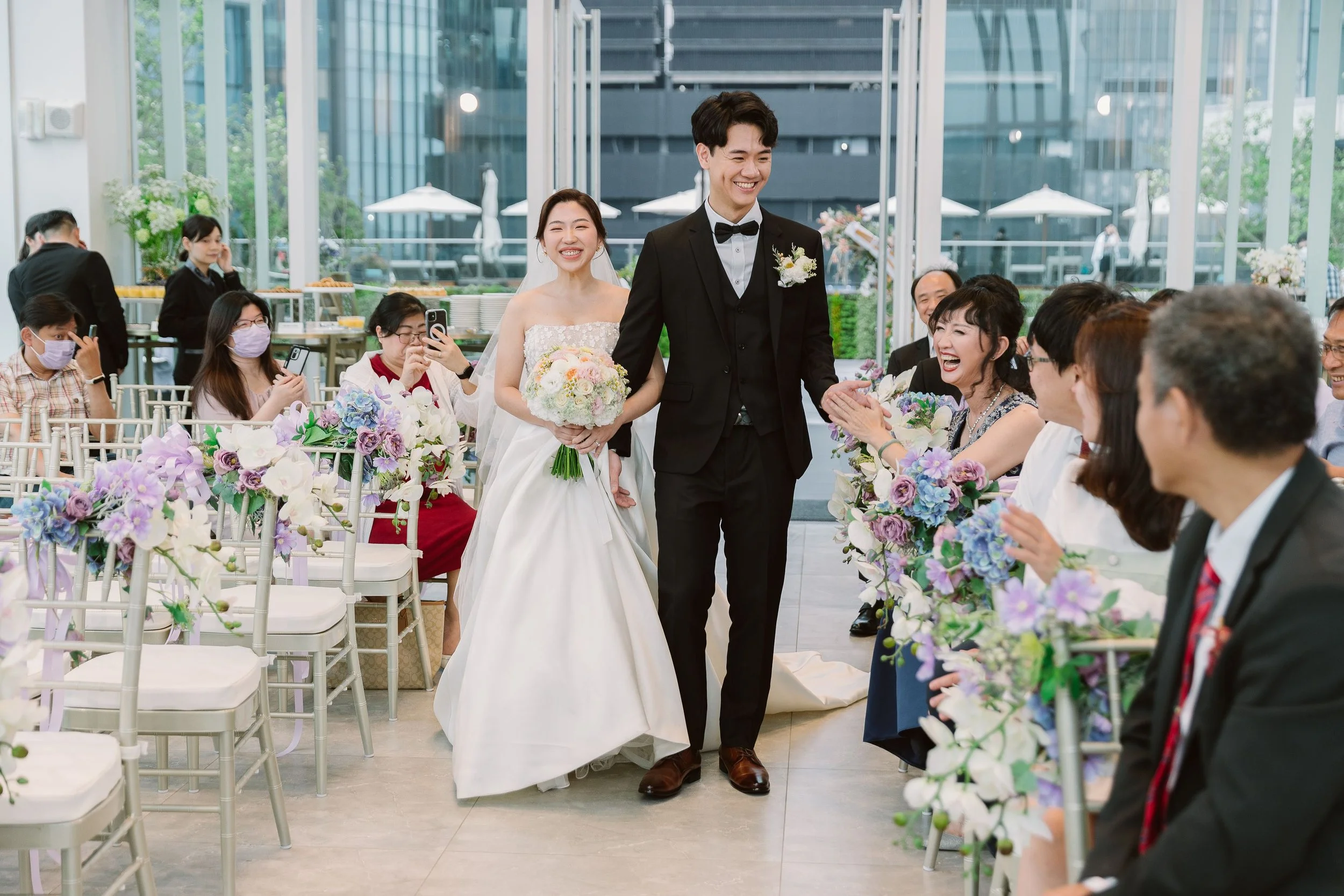 A newlywed couple walking down the aisle at a wedding reception. The bride is smiling and holding a bouquet, while the groom is smiling and holding her hand. Guests seated on either side of the aisle are smiling and taking photos. The reception is in