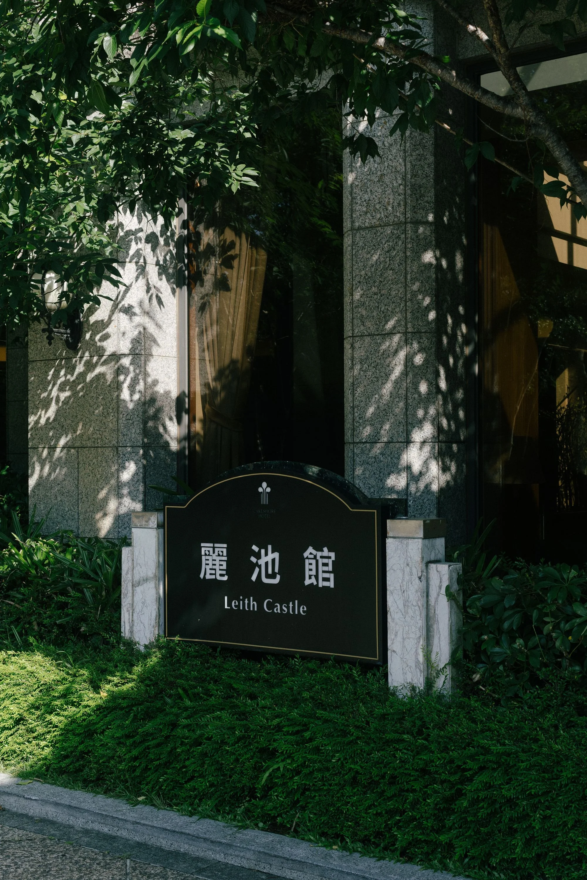 A sign with Chinese characters and the English text 'Leith Castle' on a black background, surrounded by lush green plants, with a building and window behind it.