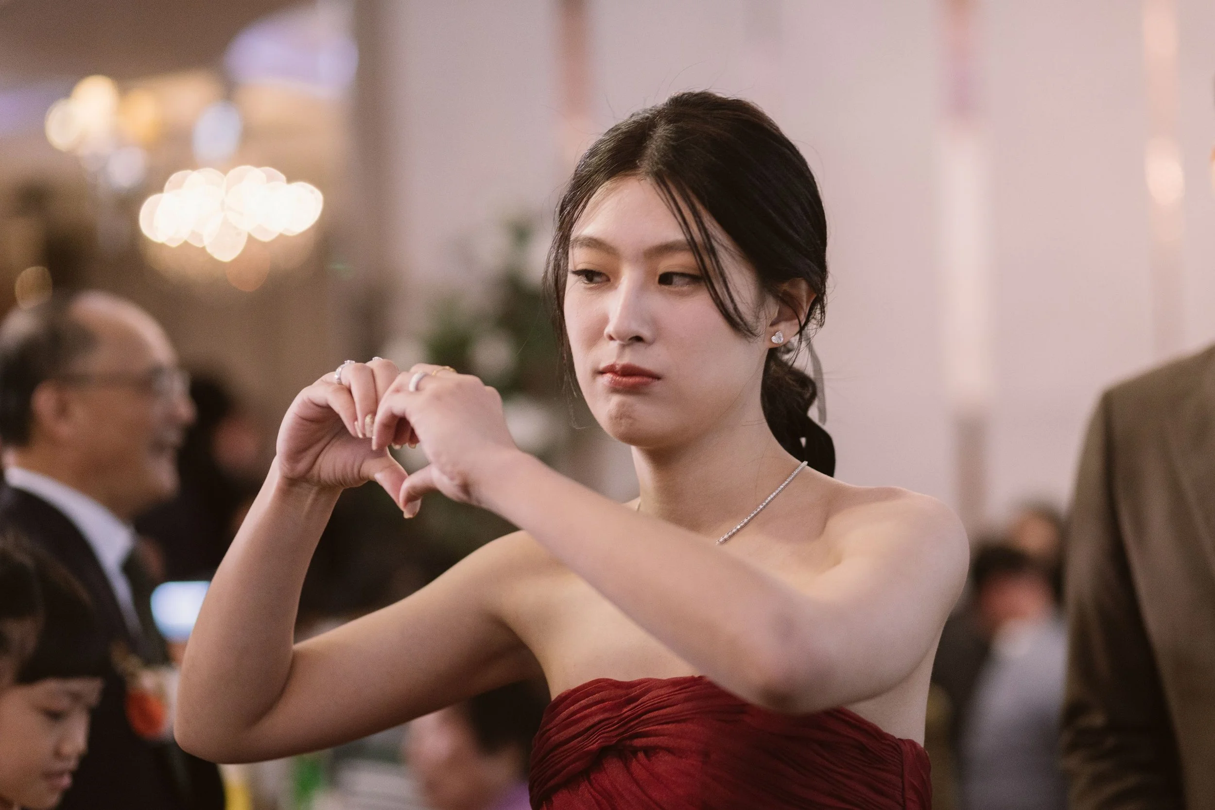 A woman wearing a strapless red dress and diamond earrings dancing with a man at a formal event. She is making a heart shape with her hands while looking to the side.