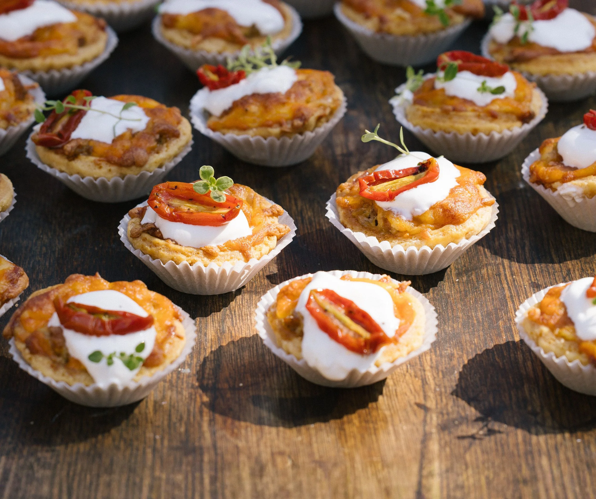 Miniature savory tartlets topped with sour cream, red peppers, shredded cheese, bacon, and microgreens arranged on a dark wooden surface.