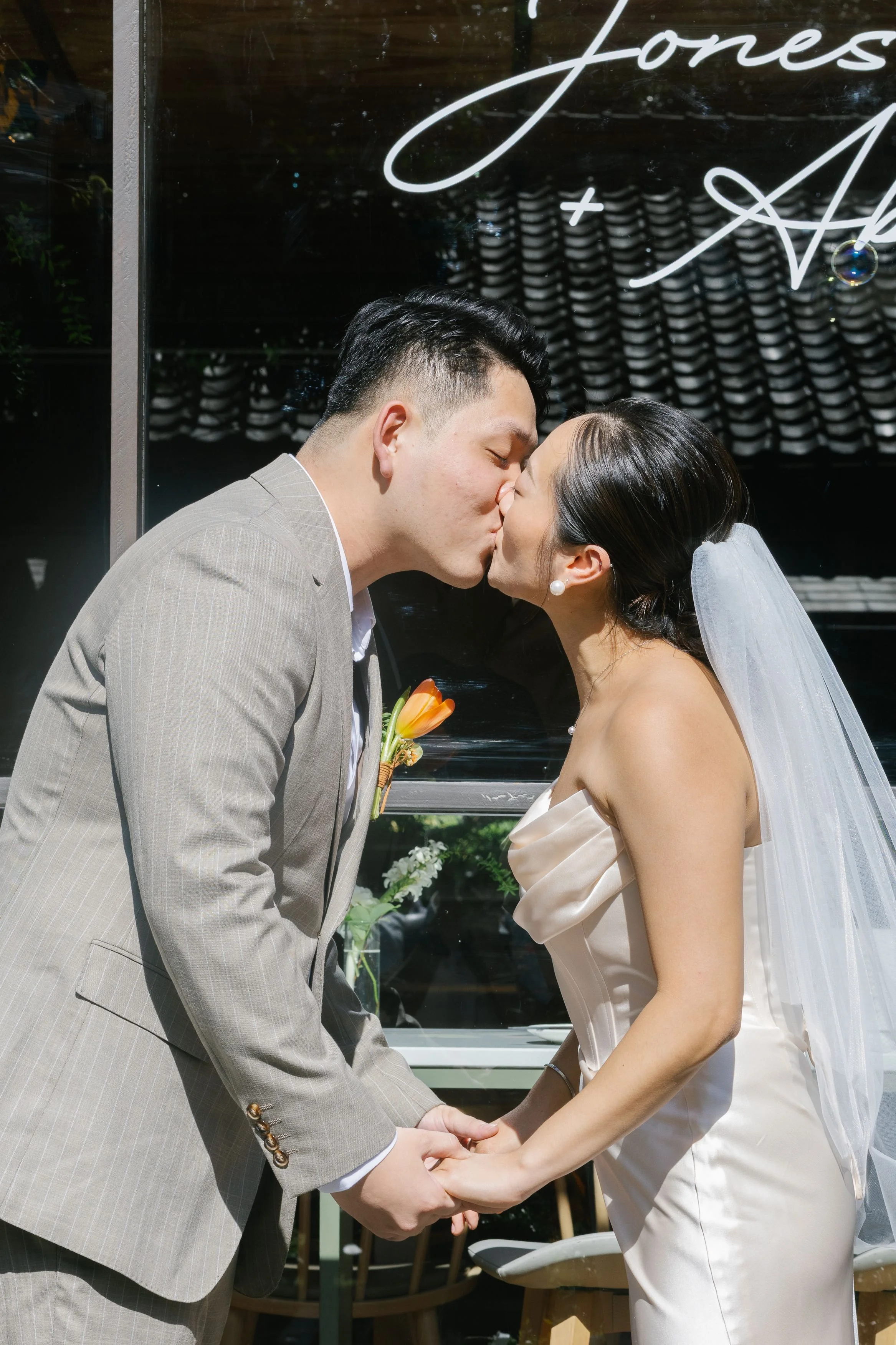 A couple is sharing a kiss and holding hands during their wedding ceremony. The groom is wearing a light gray pinstripe suit with a boutonniere, and the bride is dressed in a satin strapless wedding gown with a veil and pearl earrings. They are stand
