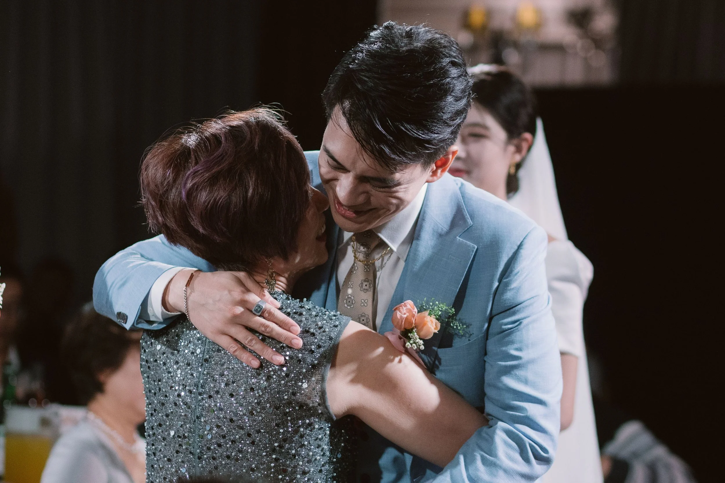 A young man in a light blue suit hugging an older woman with short reddish-brown hair, both smiling warmly at each other at a social gathering. Another woman with glasses and dark hair, wearing a white dress, is visible in the background.